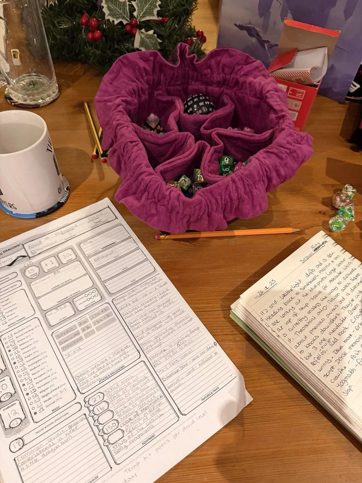 A tabletop with Dungeons & Dragons character sheets, a pink velvet drawstring bag filled with multi-colored dice, a mug, a glass jar, pencils, and some wrapped candies, with a Christmas tree in the background.