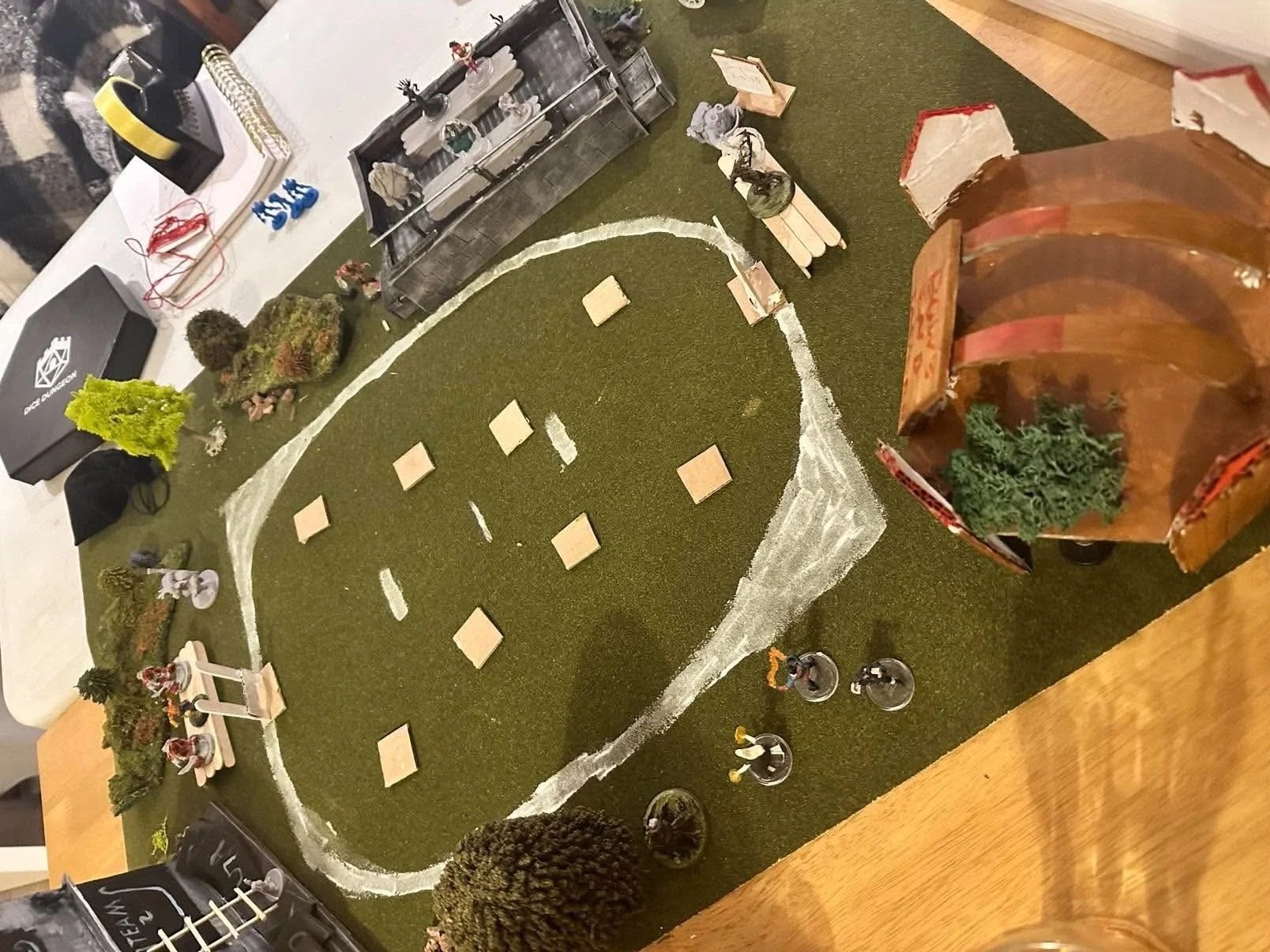 A tabletop miniature landscape features a winding road, multiple figurines of people, trees, and small structures, creating a scene for tabletop gaming or diorama display.