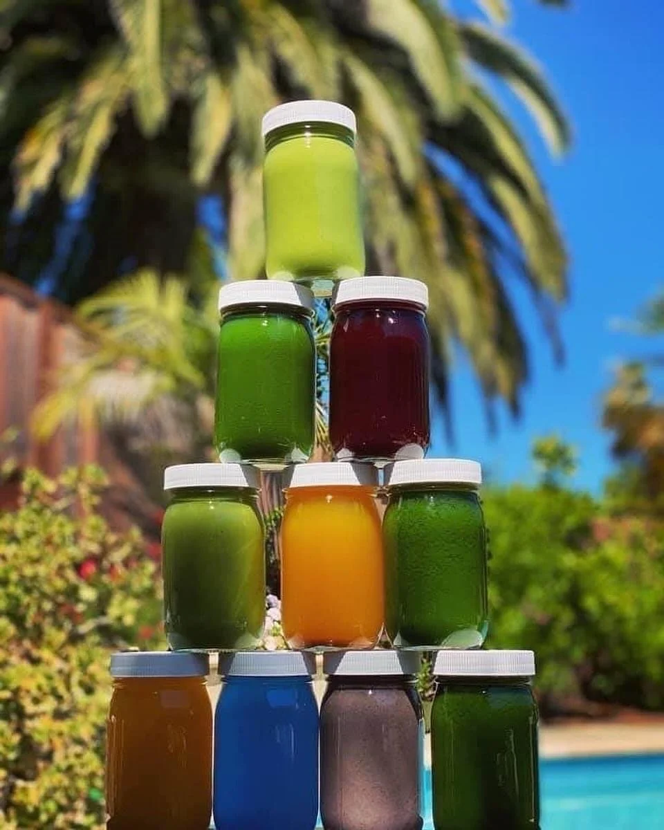 Colorful Green Your Spirit juices, elixirs, smoothies, and coconut water in glass jars stacked outdoors in front of palm trees and a swimming pool