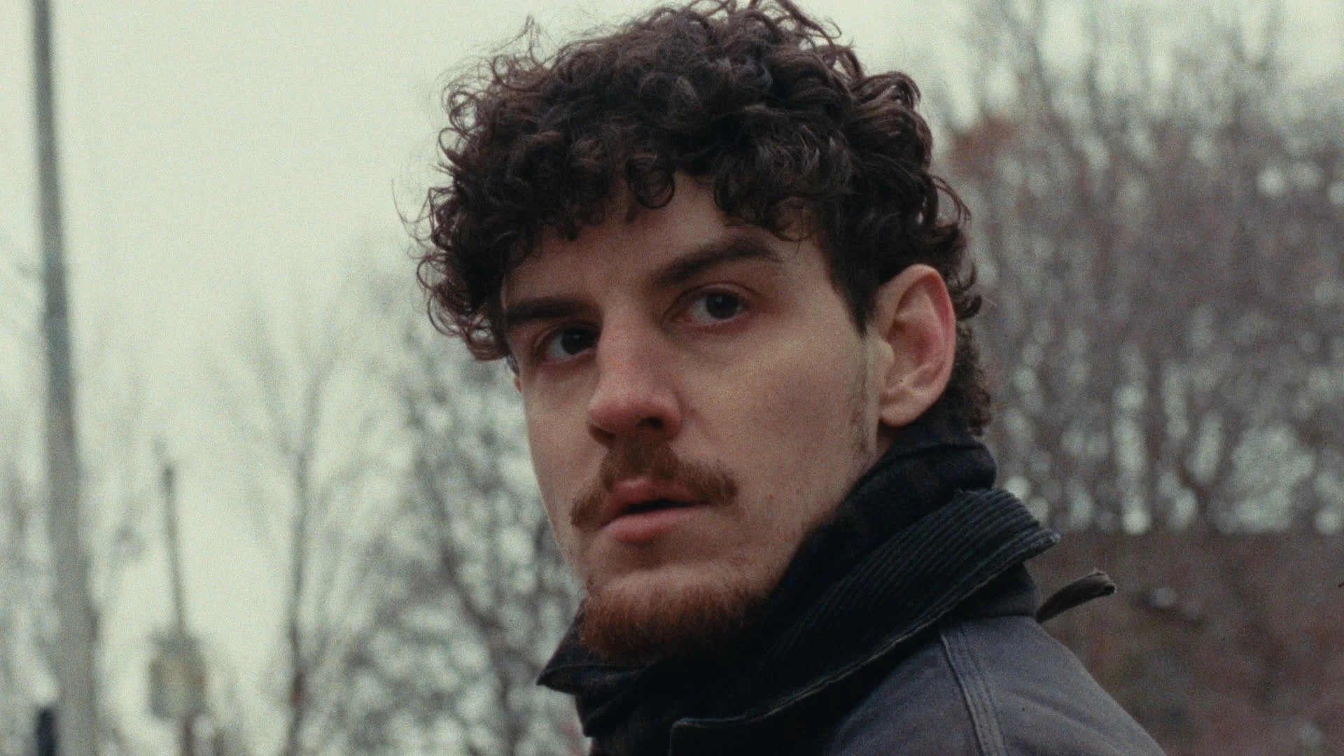 A young man with curly hair, a mustache, and a beard, looking over his shoulder outdoors with trees in the background.