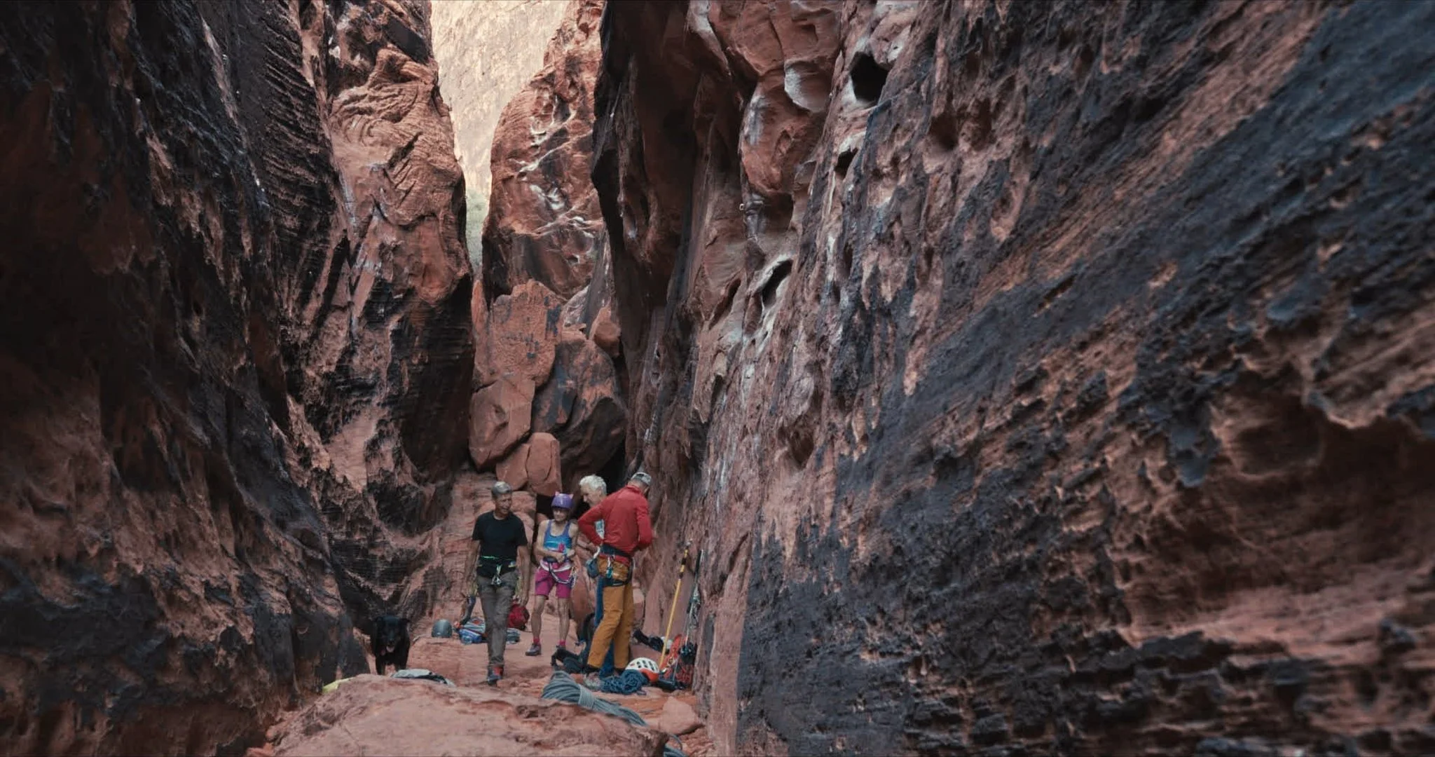 Group of people exploring a narrow, rocky canyon with red and black walls, some gear and helmets on the ground.