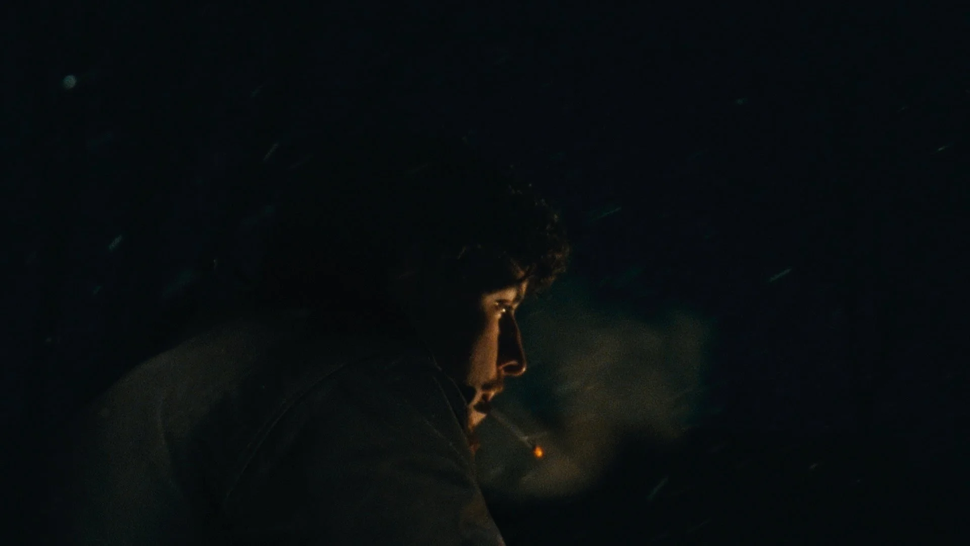 A young man with curly dark hair sitting in a dimly lit environment, with faint light illuminating his face from the side, creating a dramatic and contemplative mood.