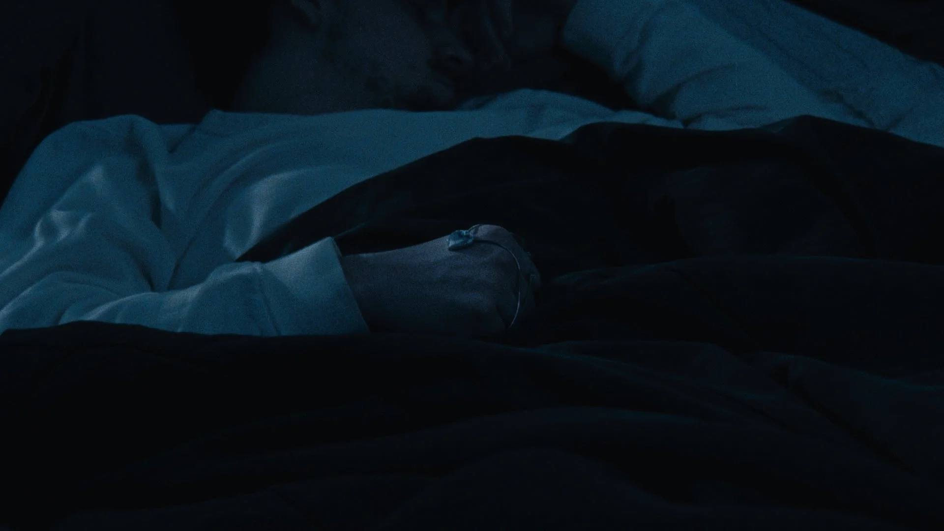 A person lying in bed with white and dark-colored blankets, wearing a grey hoodie and grey sweatpants, sleeping in the dark