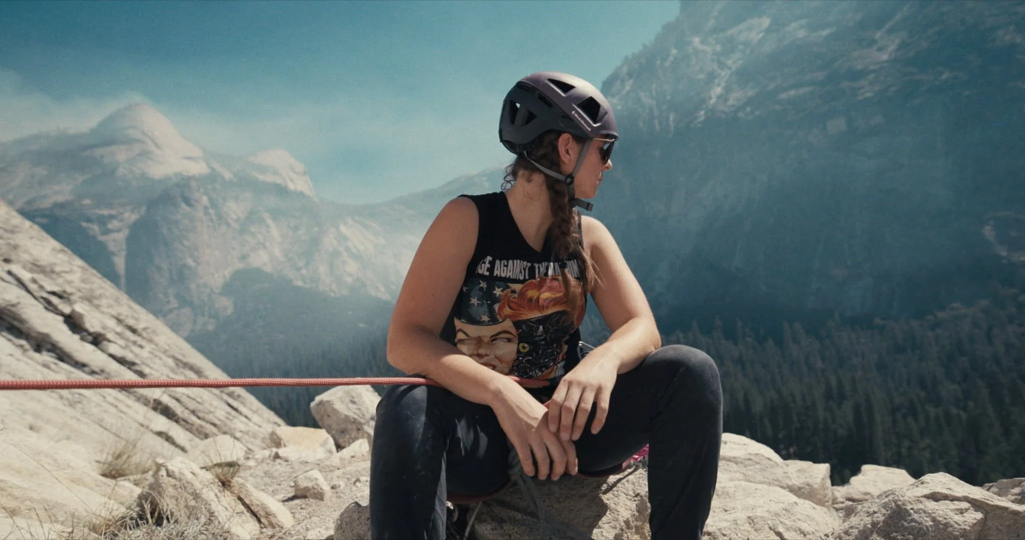 Woman wearing a bike helmet and sunglasses sitting on rocks in a mountainous landscape with snow-capped peaks in the background.