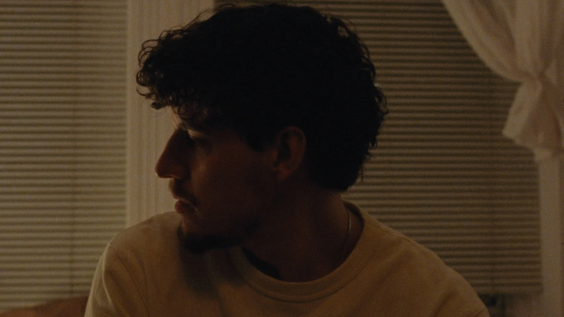 Side profile of a young man with curly hair in a dimly lit room, wearing a yellow shirt and a necklace, looking down.