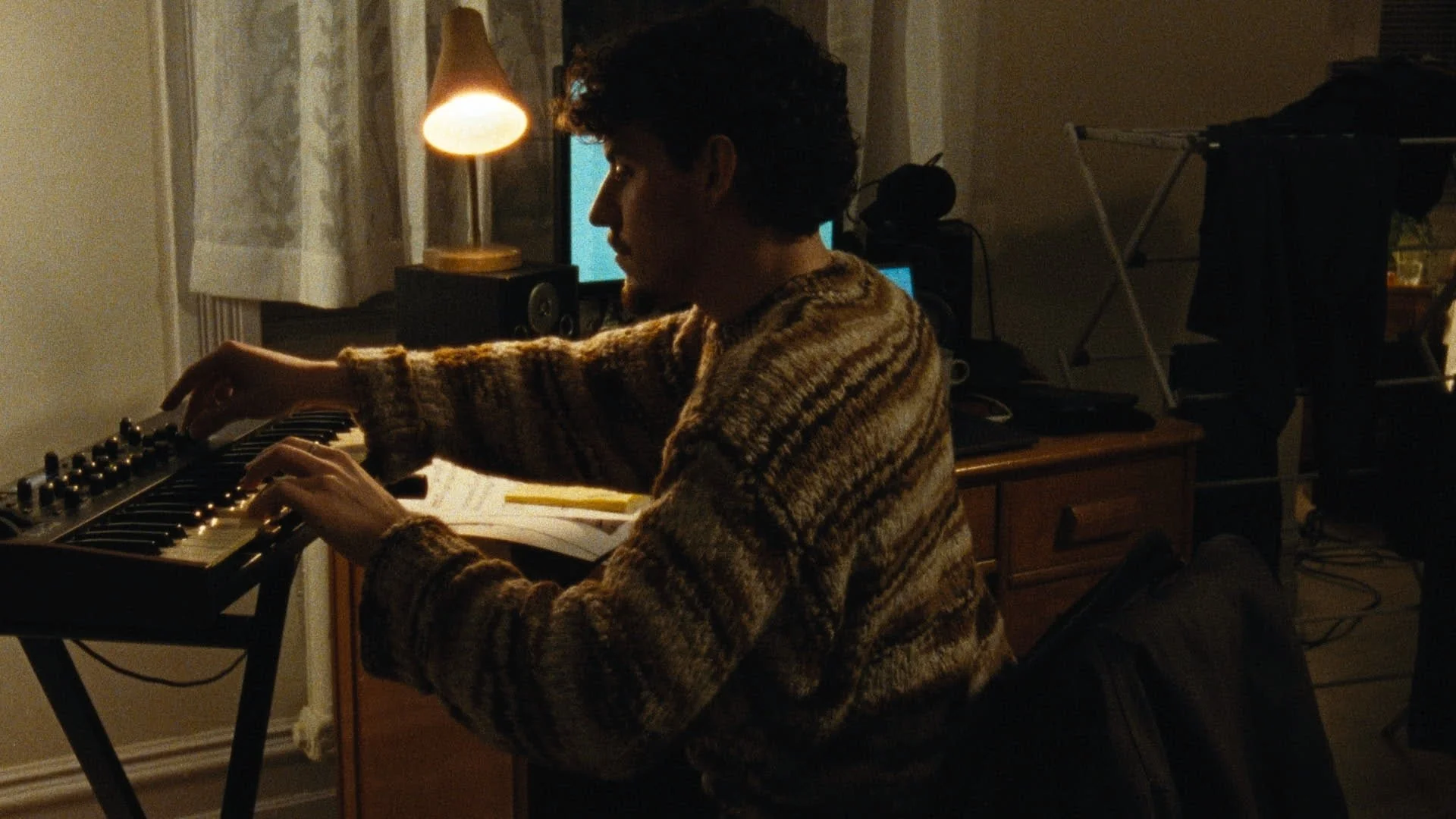 A person with dark curly hair, wearing a brown patterned sweater, playing a keyboard in a dimly lit room.
