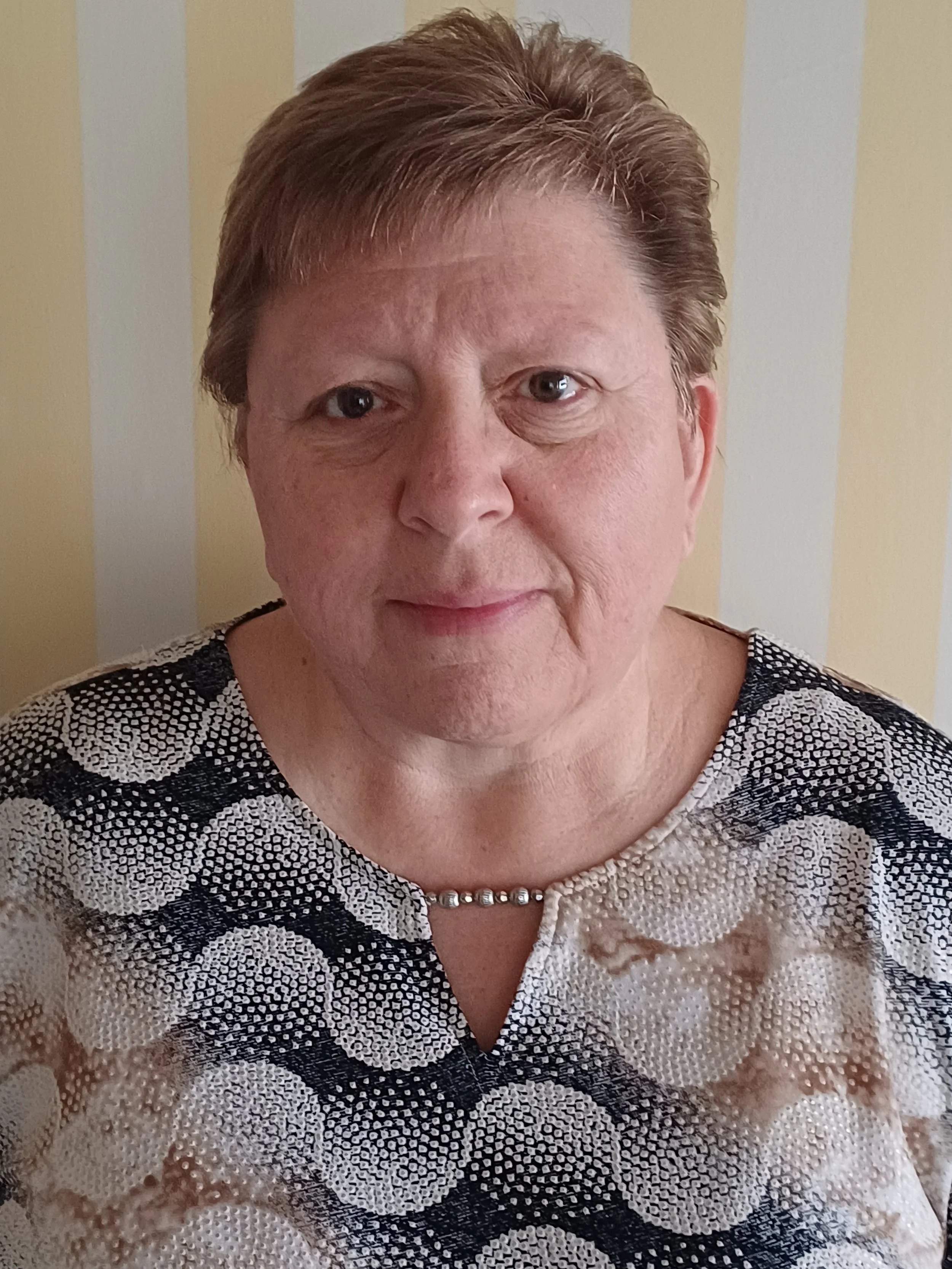 Close-up portrait of an older woman with short brown hair, wearing a patterned black-and-white blouse and necklace.