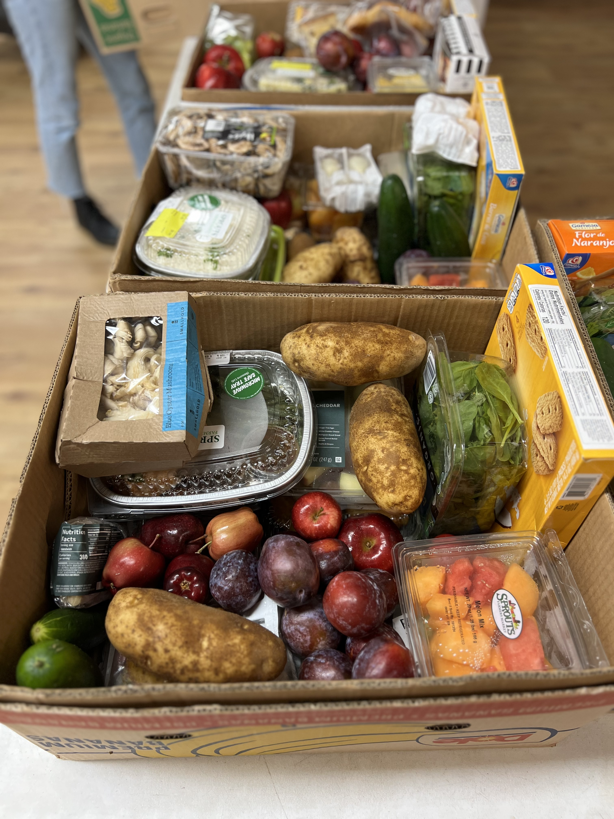 Cardboard boxes filled with assorted groceries, including potatoes, apples, plums, mushrooms, leafy greens, cucumbers, tomatoes, cheese, crackers, and prepared foods.