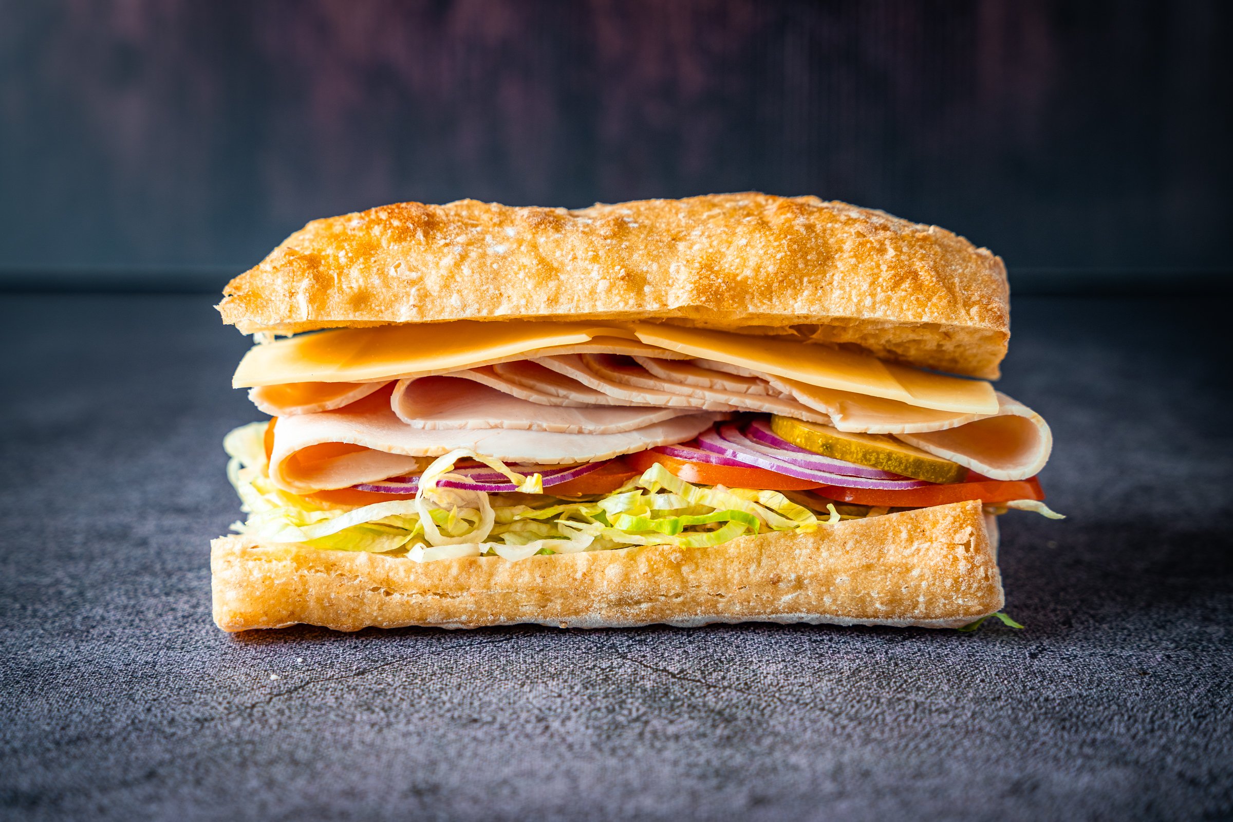 Close-up of a sandwich with sliced turkey, cheese, lettuce, tomato, and red onion on whole wheat bread, placed on a dark textured surface.