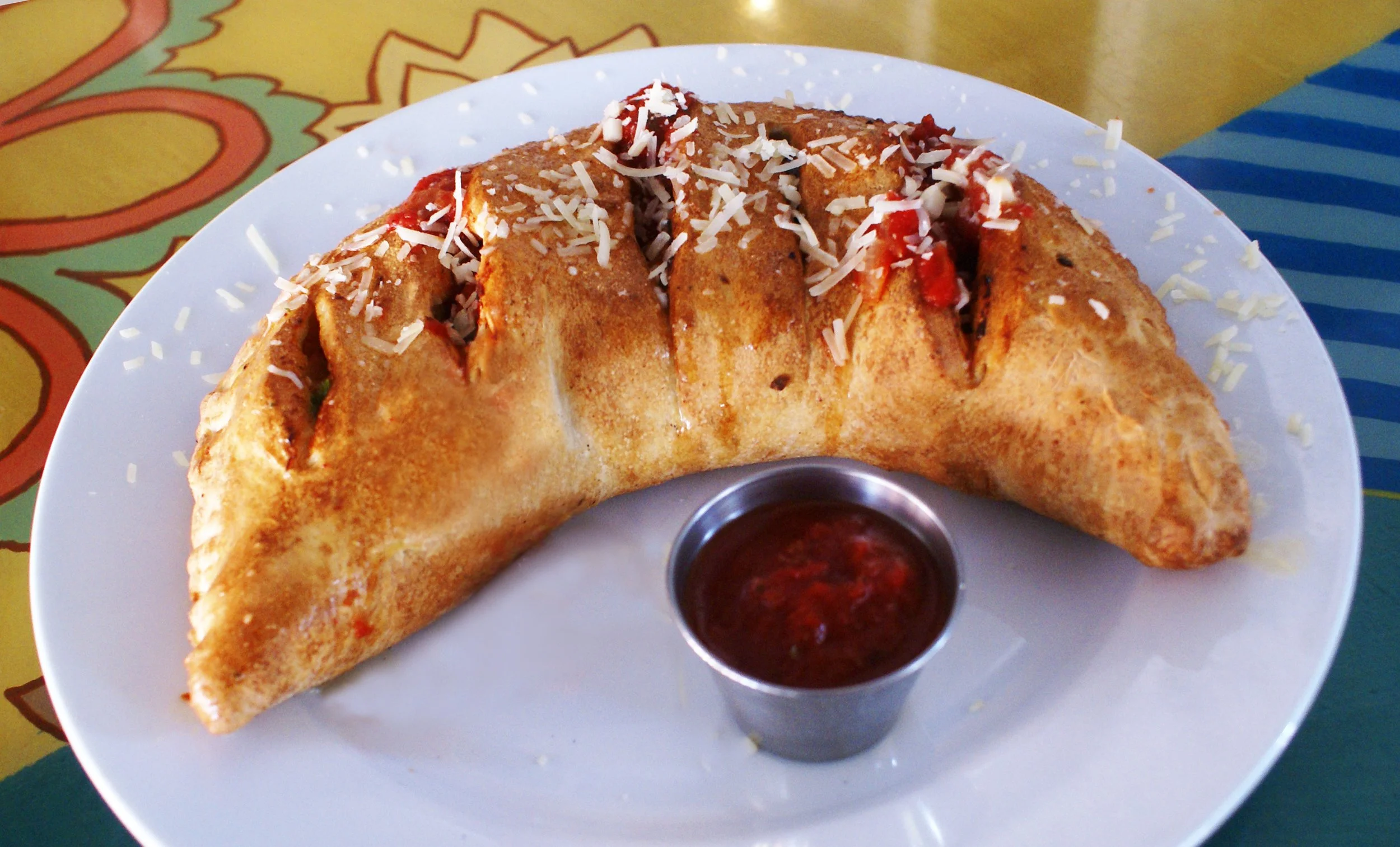 A white plate with two slices of chili and cheese stuffed pastry, topped with grated cheese and served with a small container of red sauce.