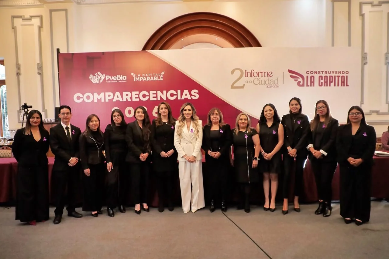 Grupo de personas vestidas formalmente, algunas con trajes negros y otras con ropa clara, de pie frente a un cartel en un evento oficial en Puebla, México, con fondo decorativo y logos relacionados con el gobierno de la ciudad y proyectos de desarrollo.