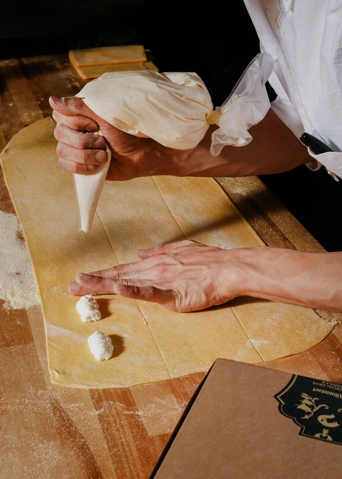 Hands shaping pasta dough on a floured wooden work surface with ricotta filling portions nearby