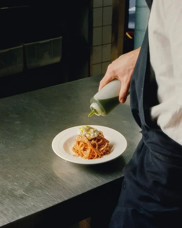Chef drizzling olive oil over a plated serving of spaghetti in a commercial kitchen