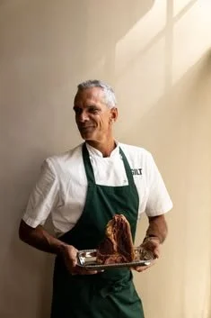 Smiling older chef in a green apron holding a roasted turkey on a serving platter