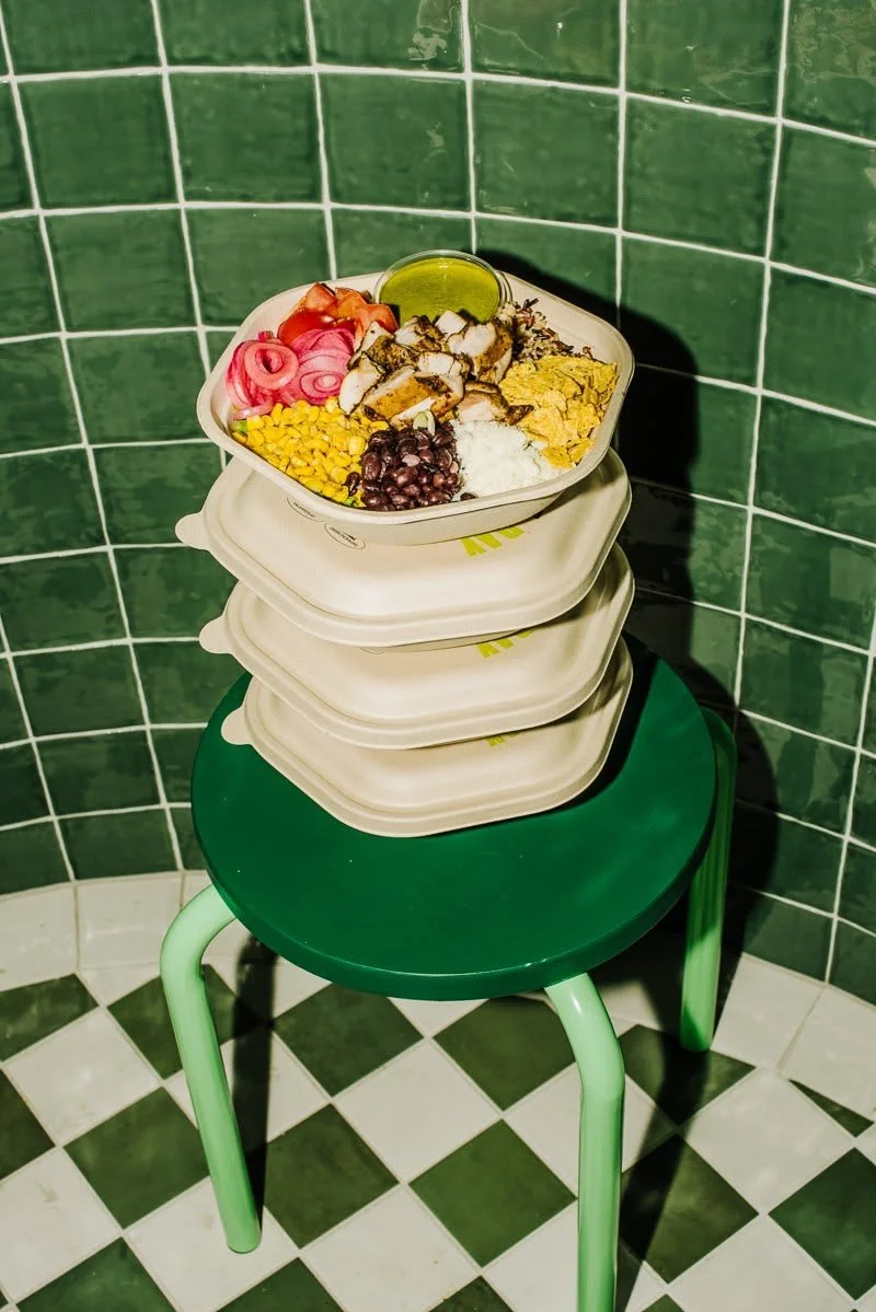 Takeout bowl of rice, grilled chicken, black beans, corn, pickled onions, tomatoes, and tortilla chips stacked on a green stool