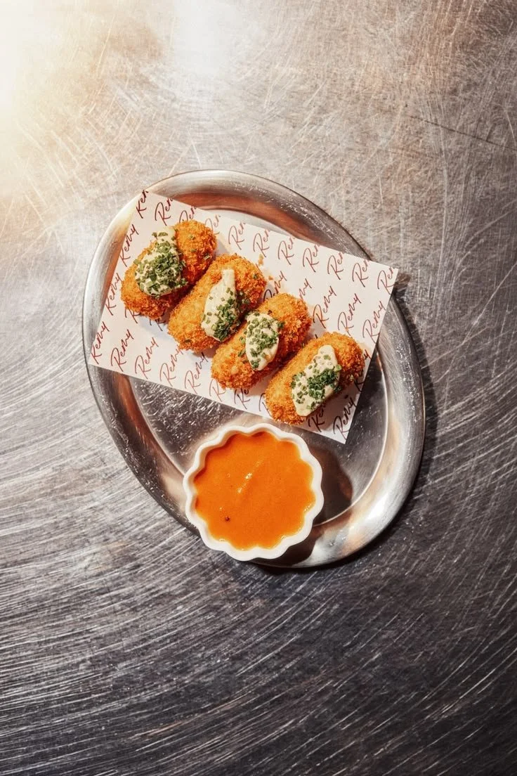 Four crispy breaded croquettes topped with herbs and creamy sauce on a branded paper-lined metal tray, served with orange dipping sauce.
