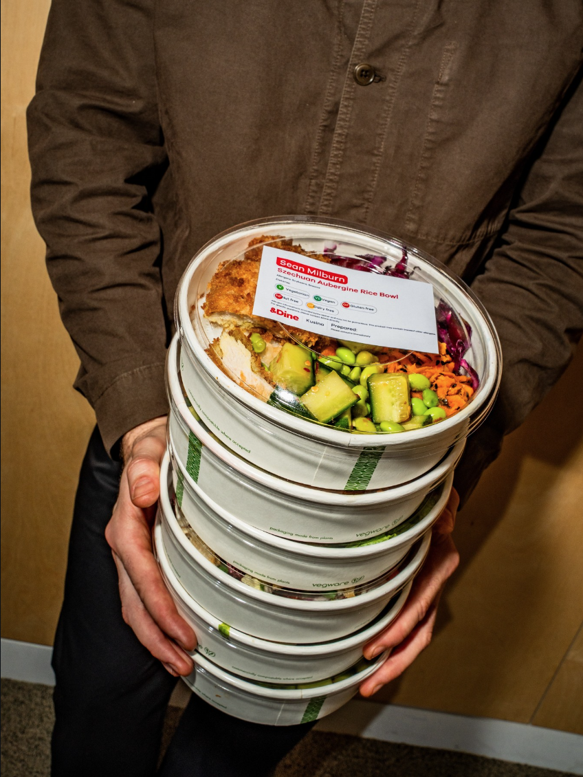 Person holding a stack of packaged vegan meal bowls, including a Szechuan aubergine rice bowl with vegetables.