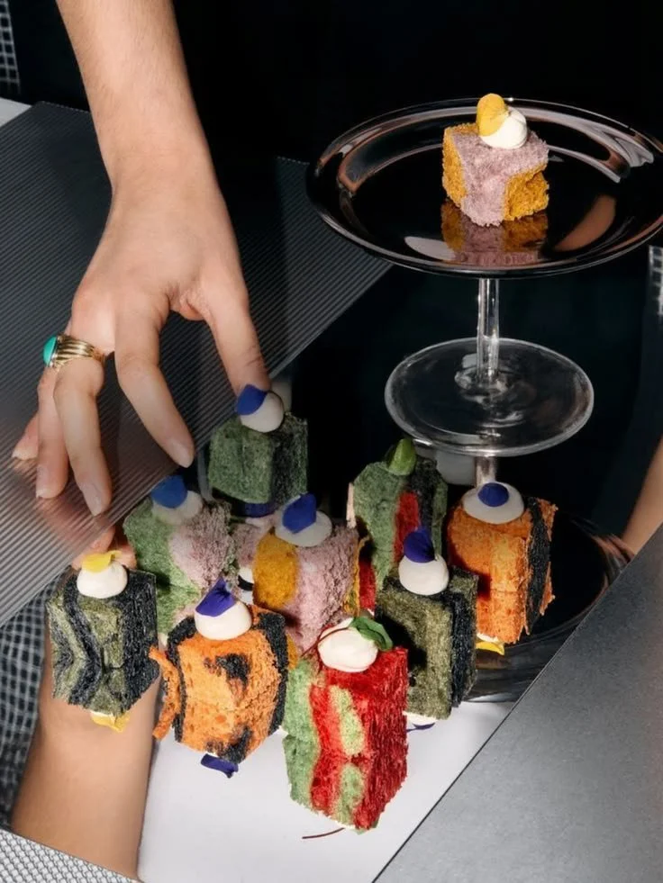 Colorful cube-shaped desserts with whipped cream and fruit garnishes on a black plate and serving glass