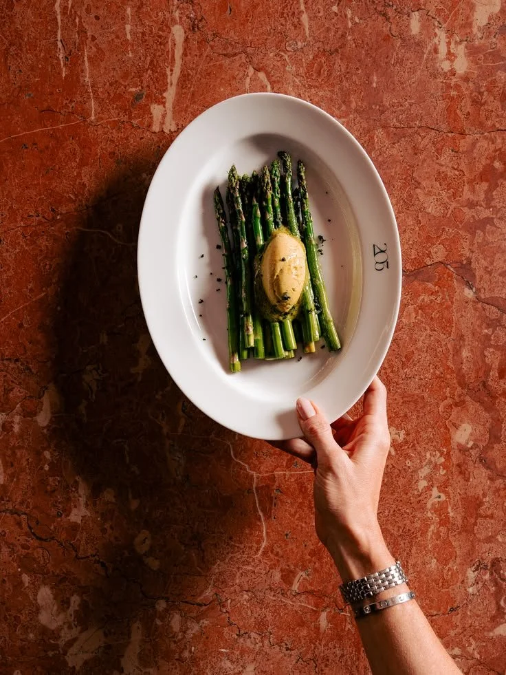 White plate of grilled asparagus topped with a poached egg and black pepper on a red marble table