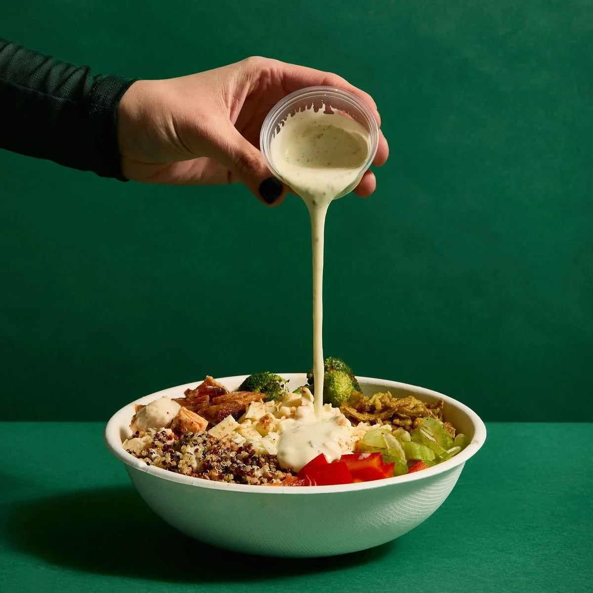 Hand pouring creamy dressing over a colorful grain and vegetable bowl