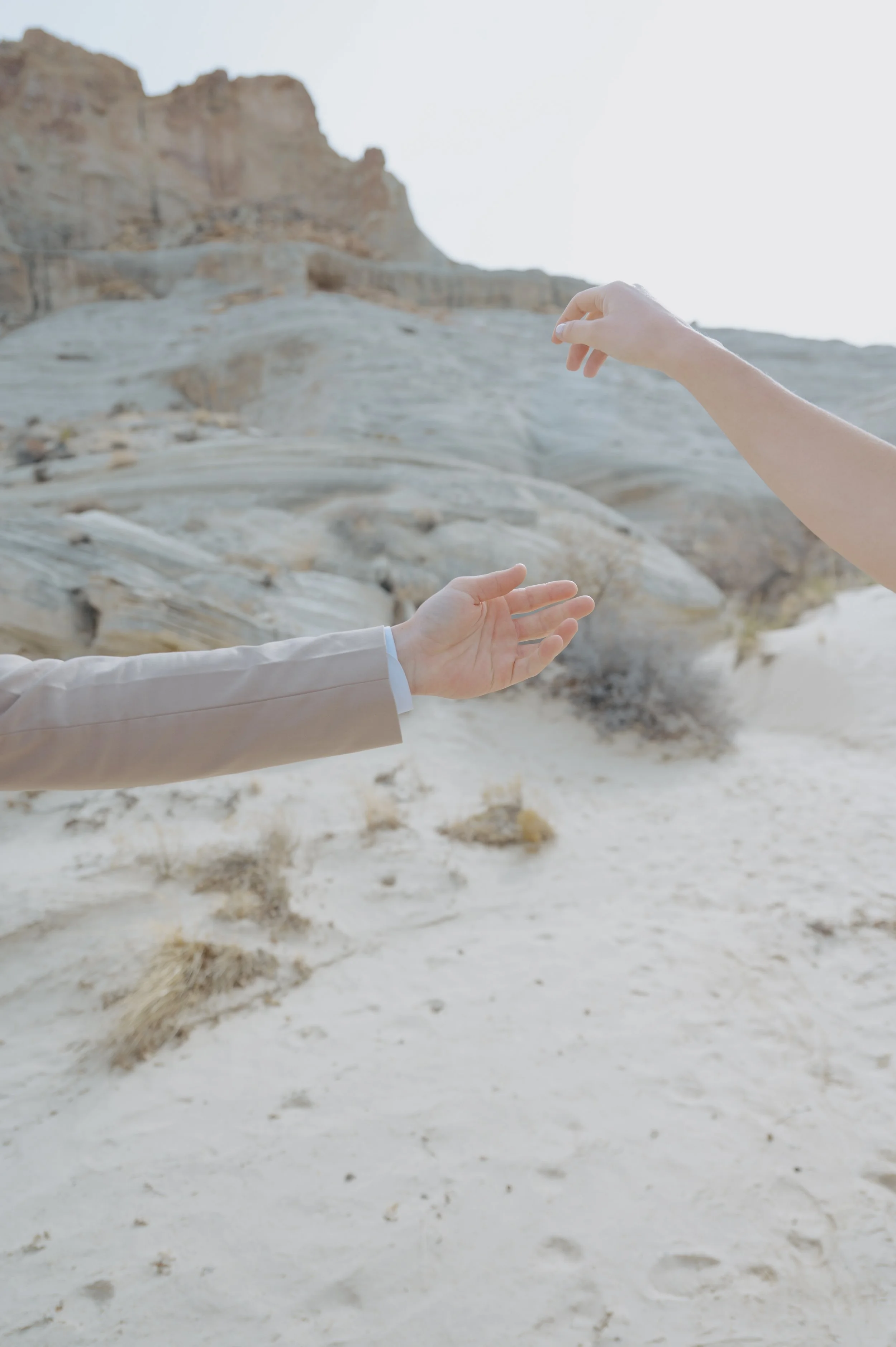 Two hands reaching toward each other across pale desert rock and sand.