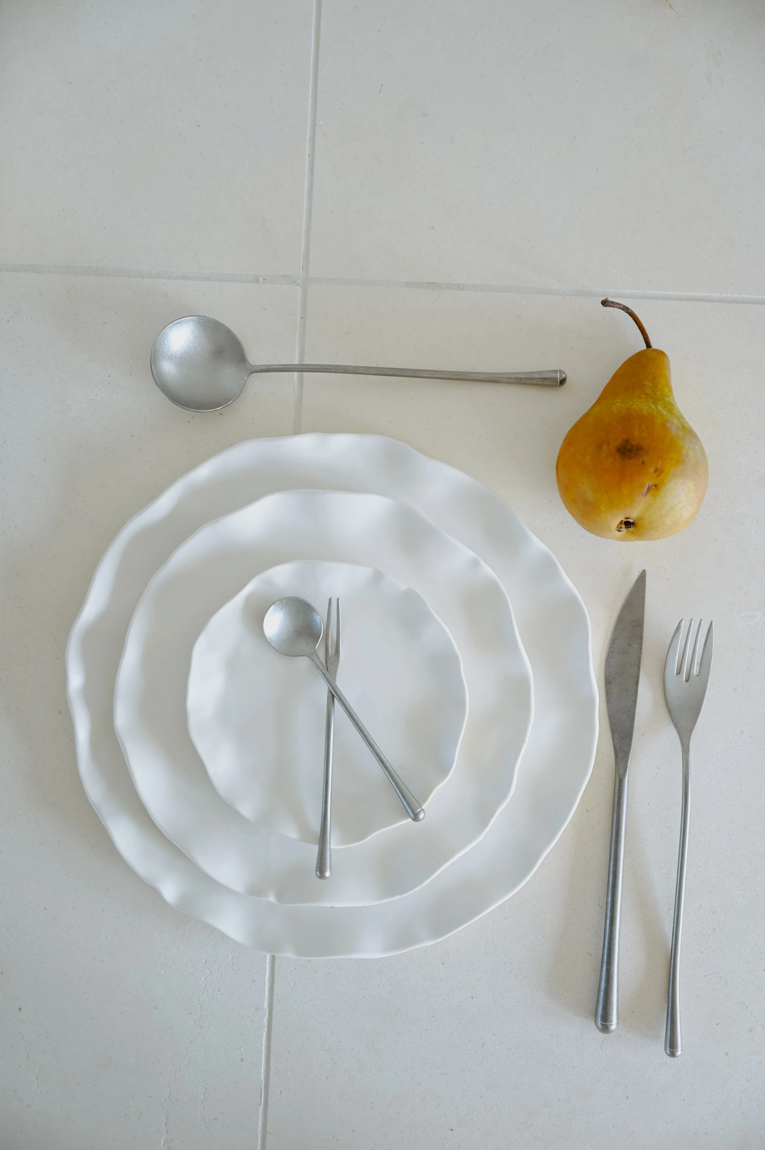 An overhead place setting with layered white plates, silver flatware and a pear.