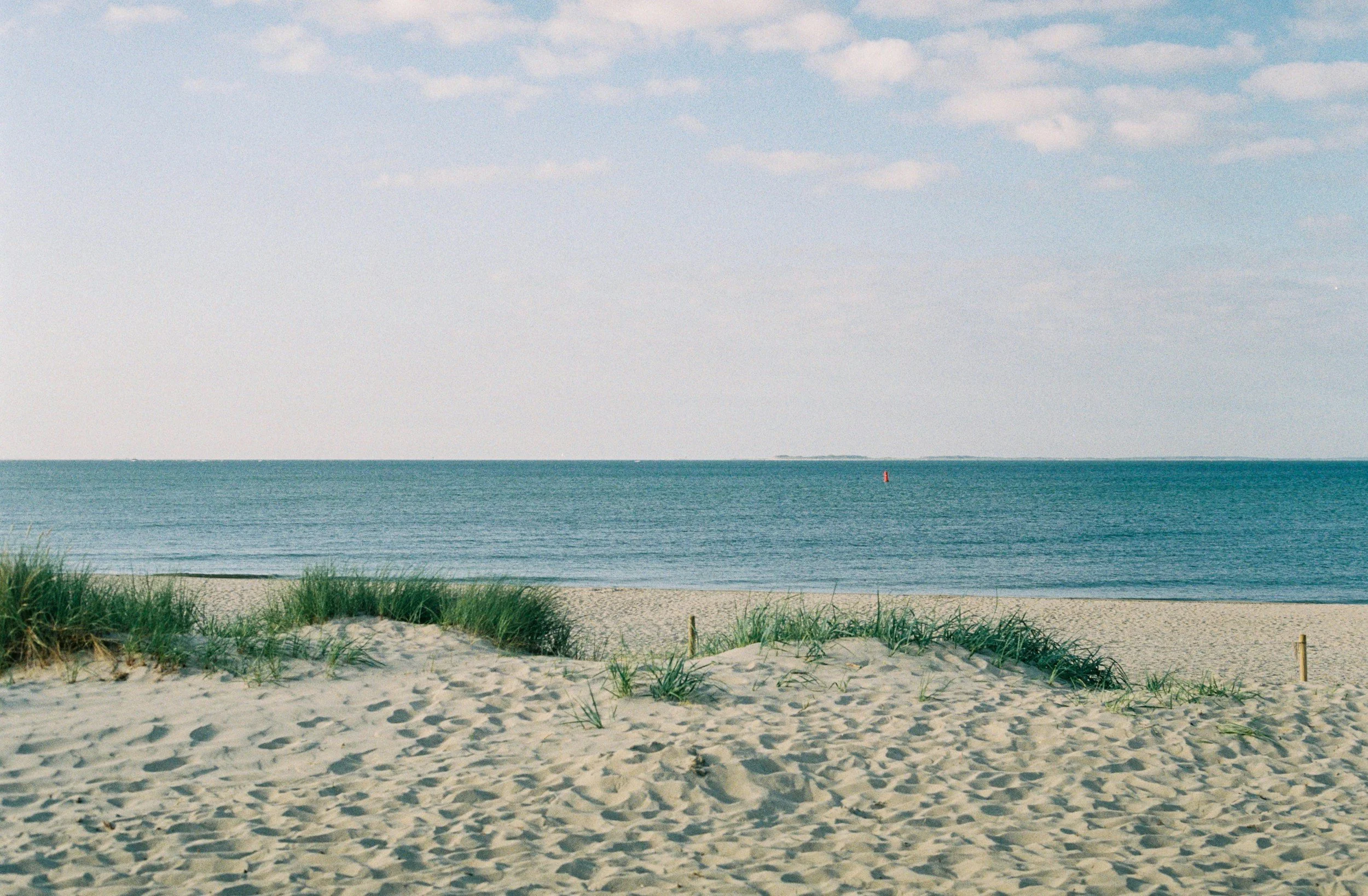 Sandy beach with dune grass, calm ocean, red buoy, and cloudy blue sky