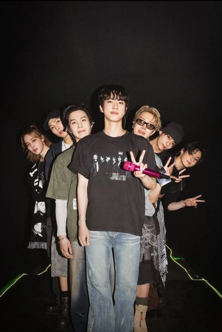 Seven performers pose together on a dark stage, with the front person holding a bright pink microphone and making a peace sign.