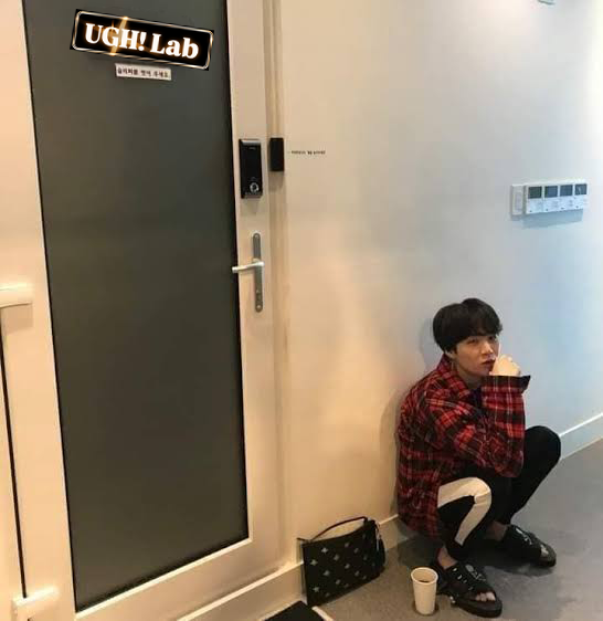 Person in a red plaid shirt sitting on the floor beside the UGH! Lab door, with a paper cup and black bag nearby