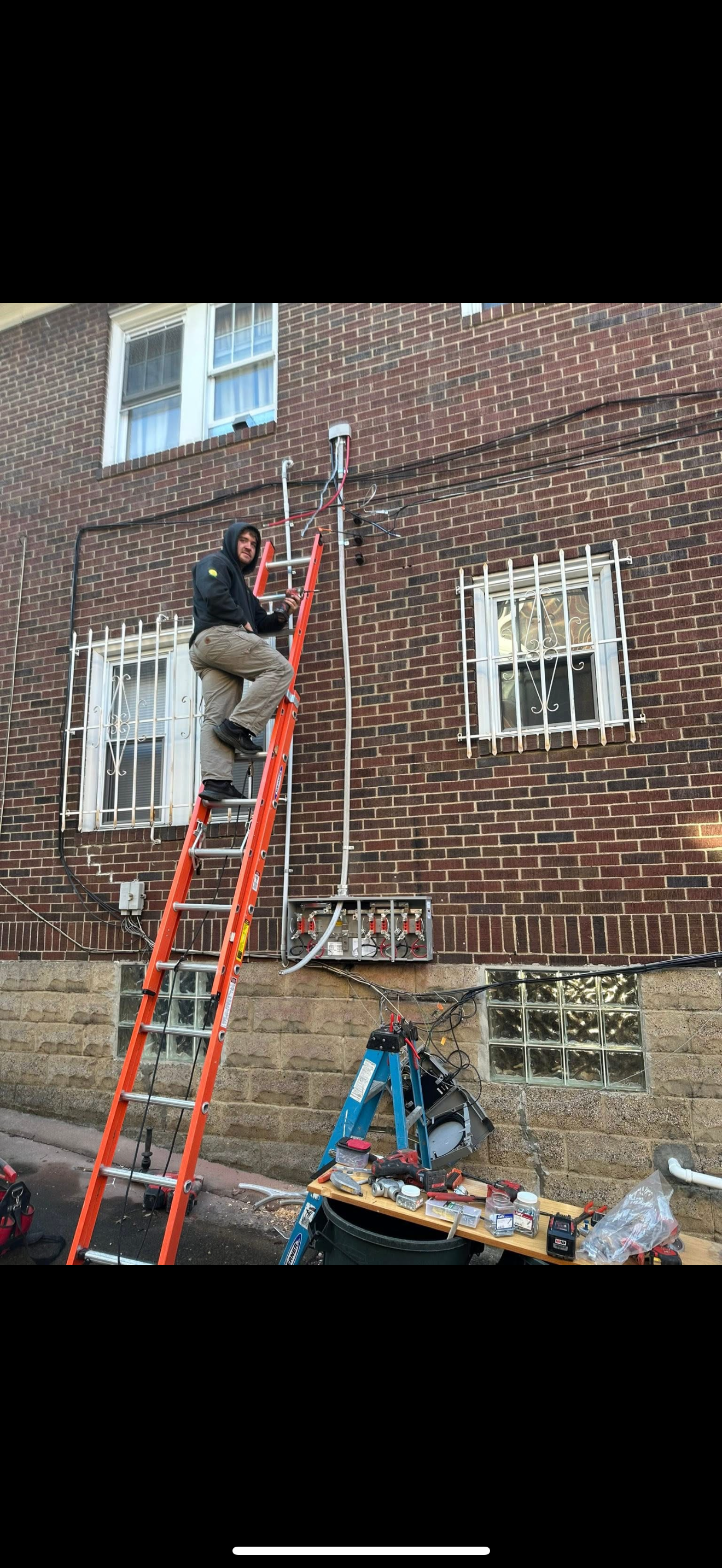 A technician working on electrical wiring on the exterior of a brick building, standing on an orange ladder, with tools and equipment nearby.
