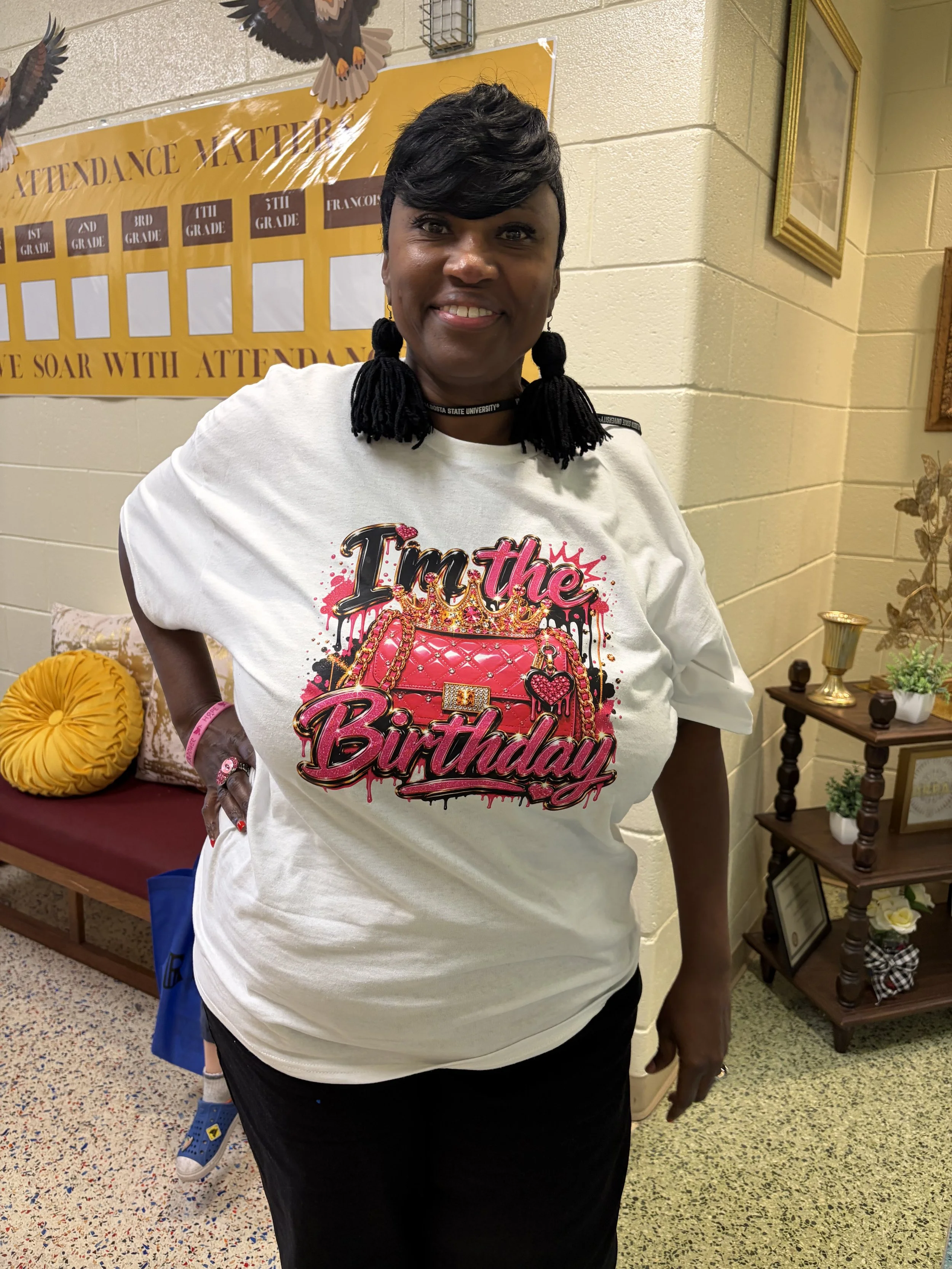 Smiling woman wearing a birthday T-shirt and tassel earrings indoors beside a school attendance chart.