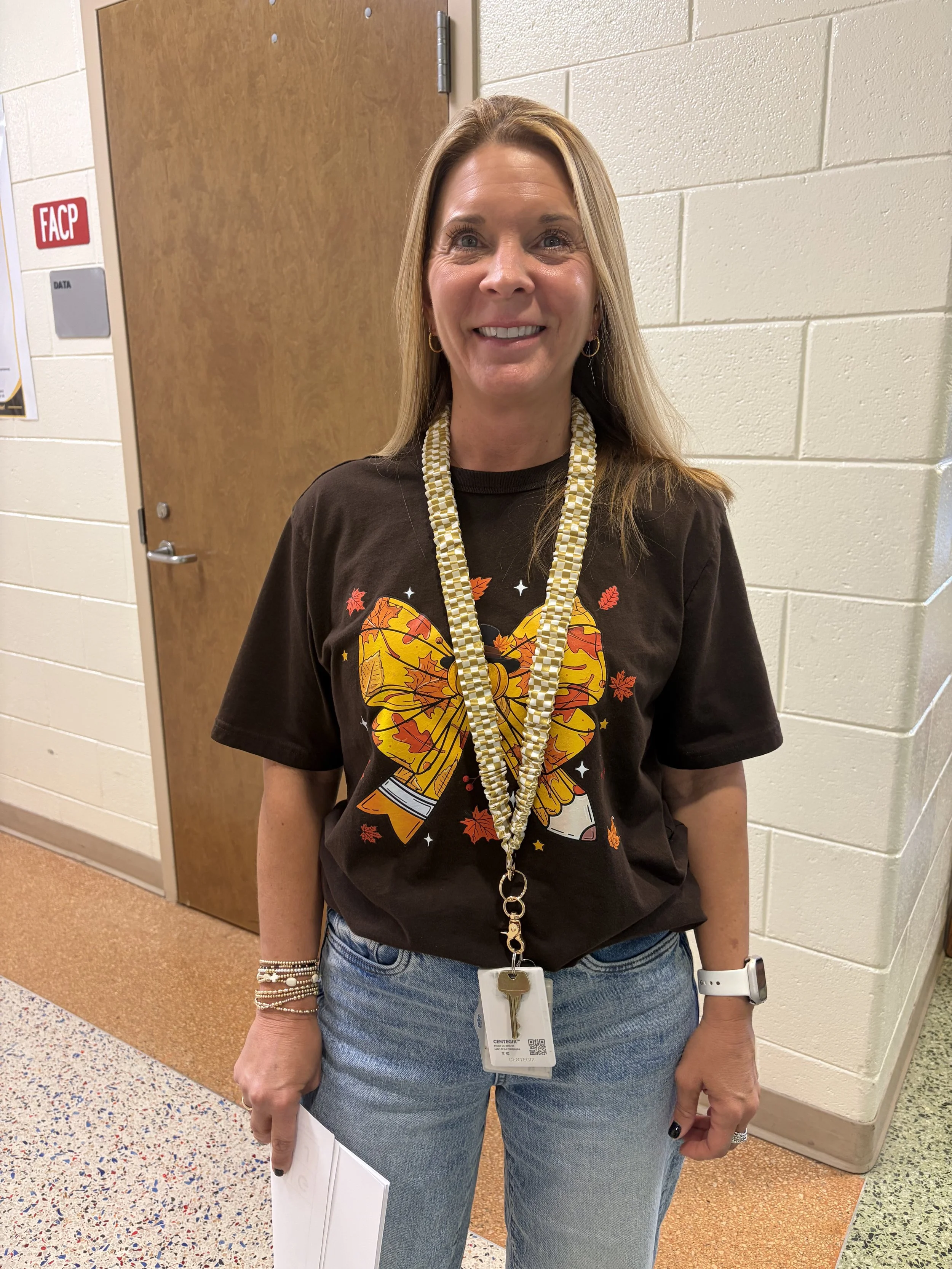 Smiling woman wearing a fall-themed black T-shirt, lanyard, and ID badge in a school hallway.