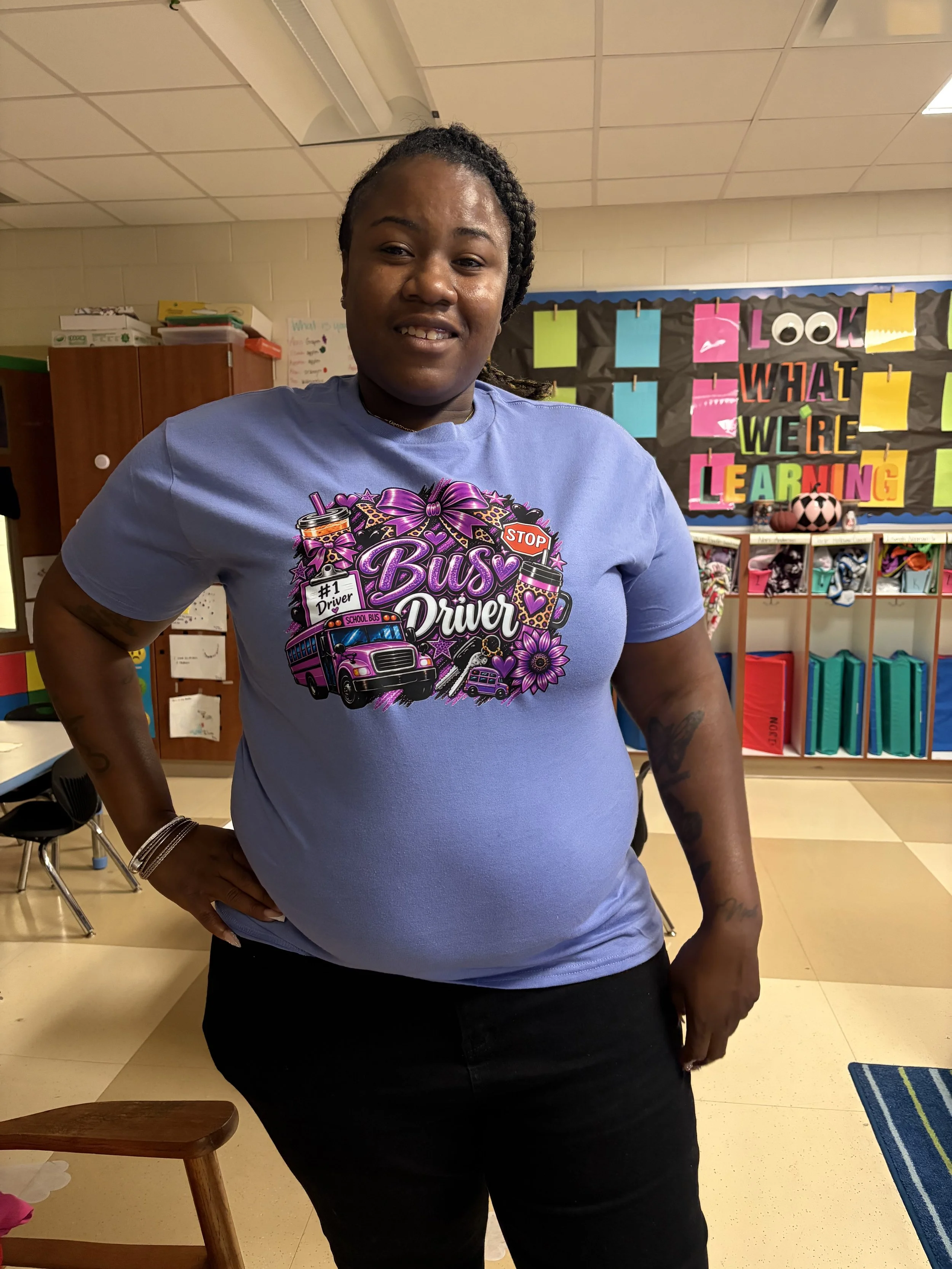 Smiling teacher in a purple school bus driver shirt standing in a colorful elementary classroom.
