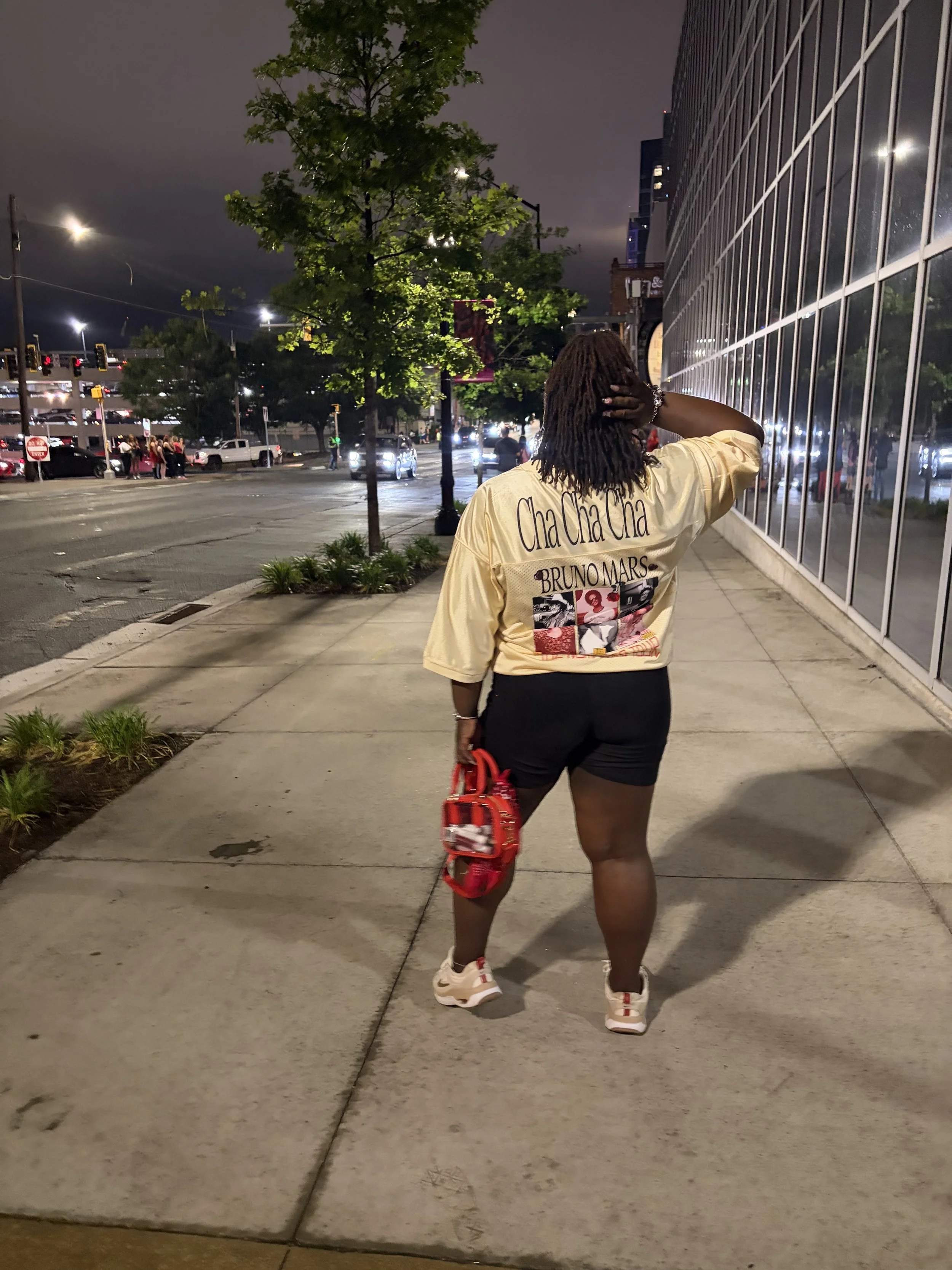 Person with long dreadlocks wearing a Bruno Mars shirt walks along a city sidewalk at night beside a glass-fronted building.