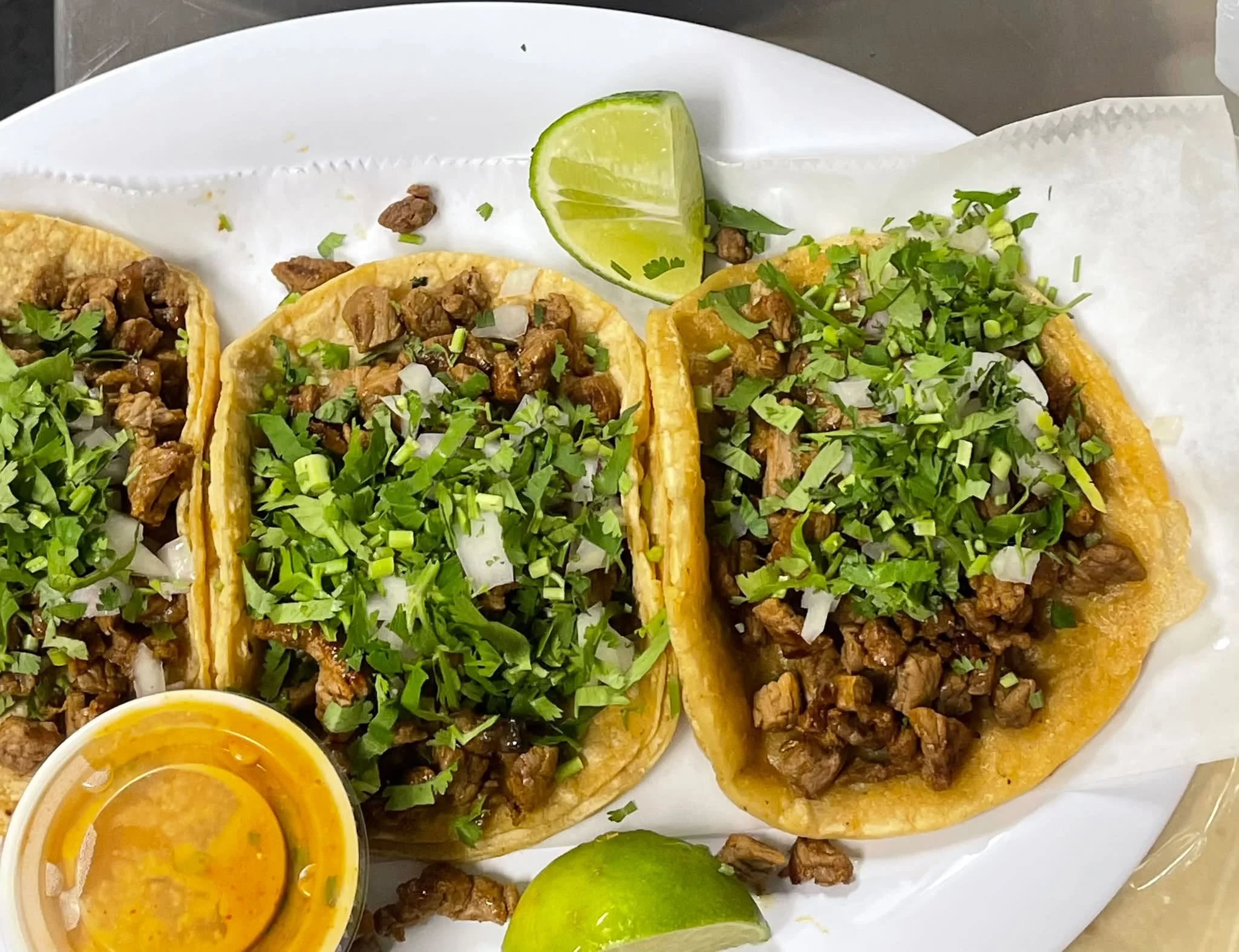 Close-up of three carne asada street tacos topped with chopped cilantro and onions, with lime wedges and a small container of orange sauce on a white plate.