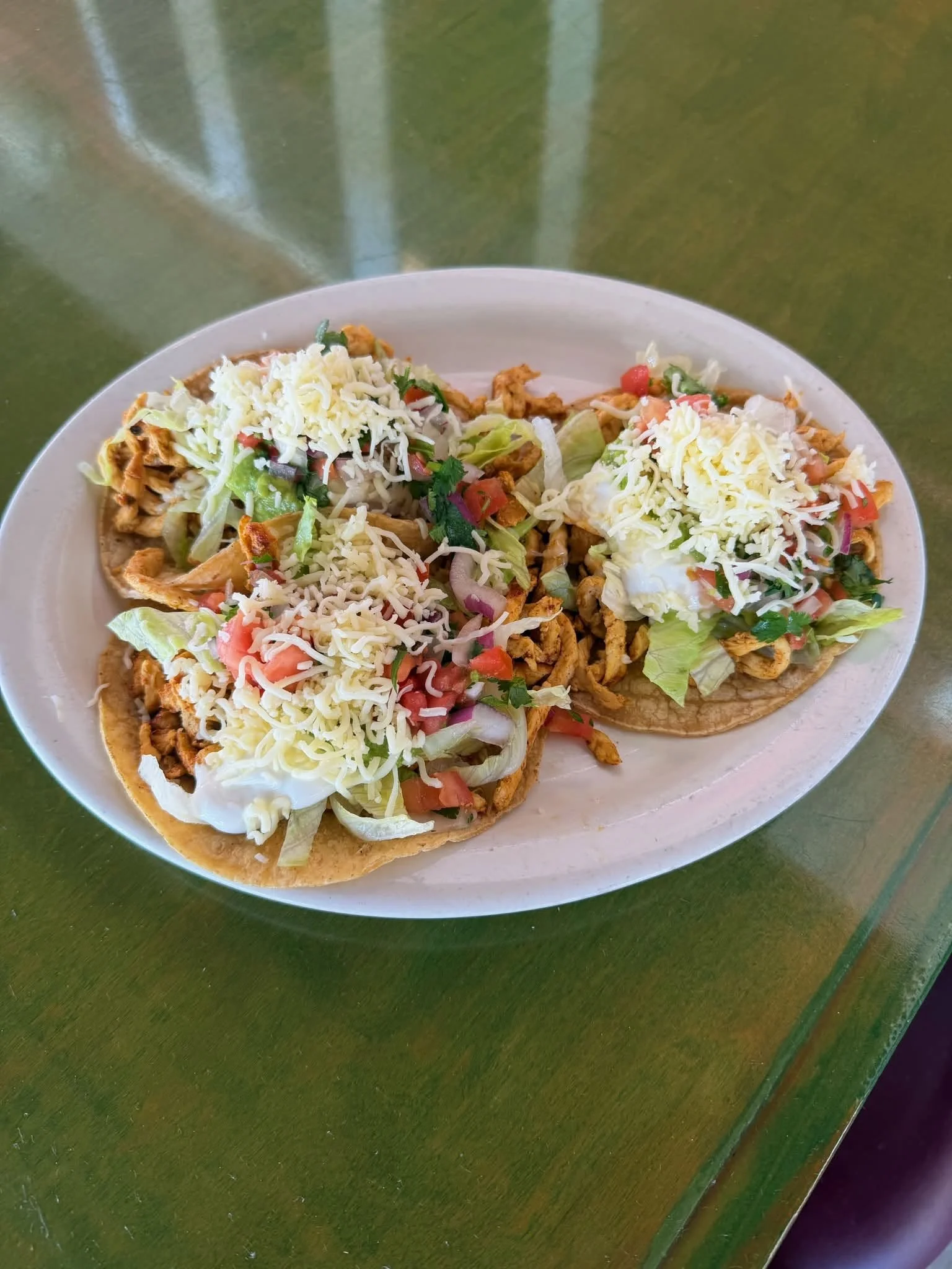 Close-up of two Mexican tacos topped with shredded cheese, lettuce, tomato, and chopped cilantro, served on a white plate on a green table.