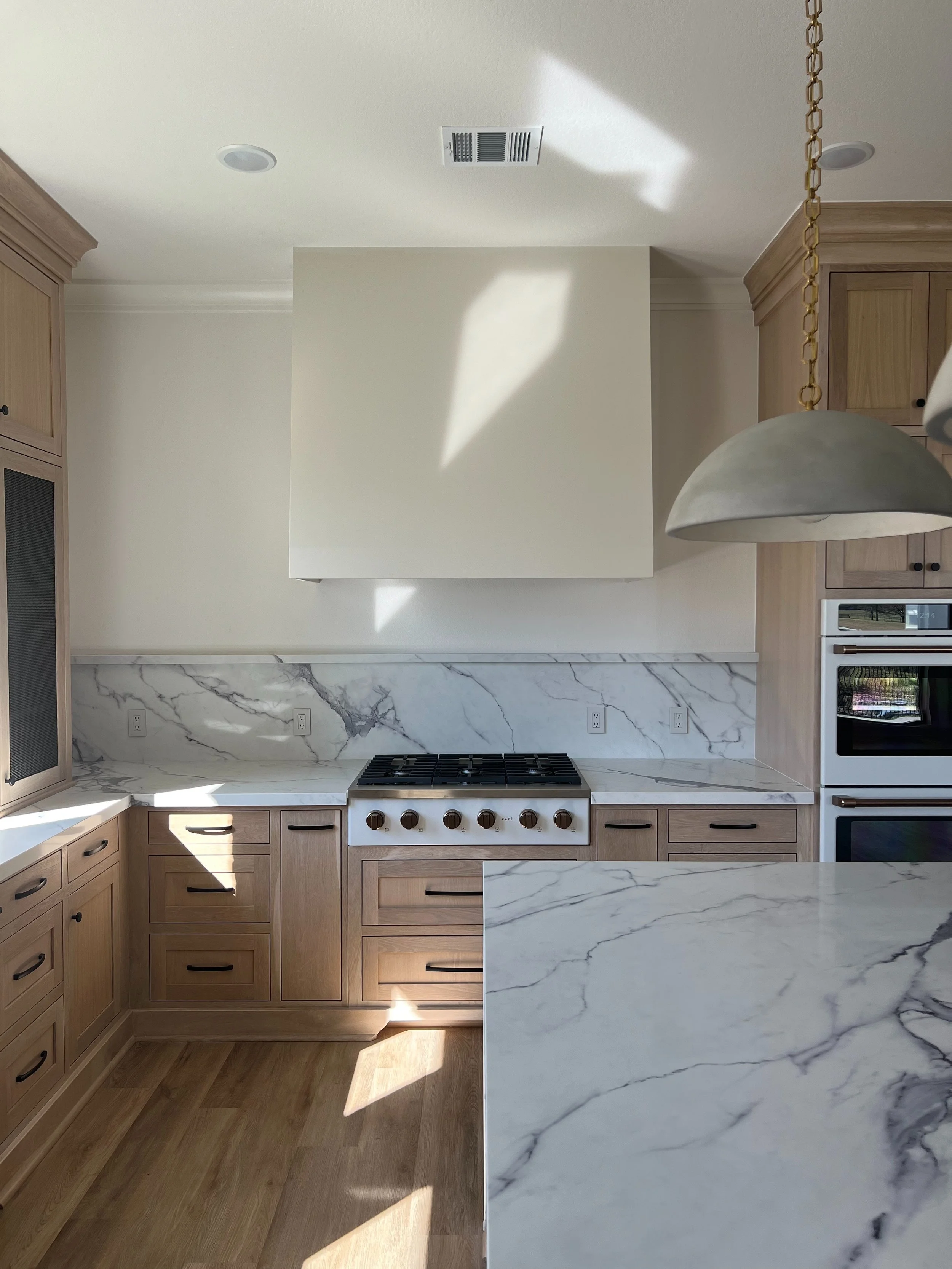 Modern kitchen with wooden cabinets, marble countertops, a stove, and a hanging light fixture.