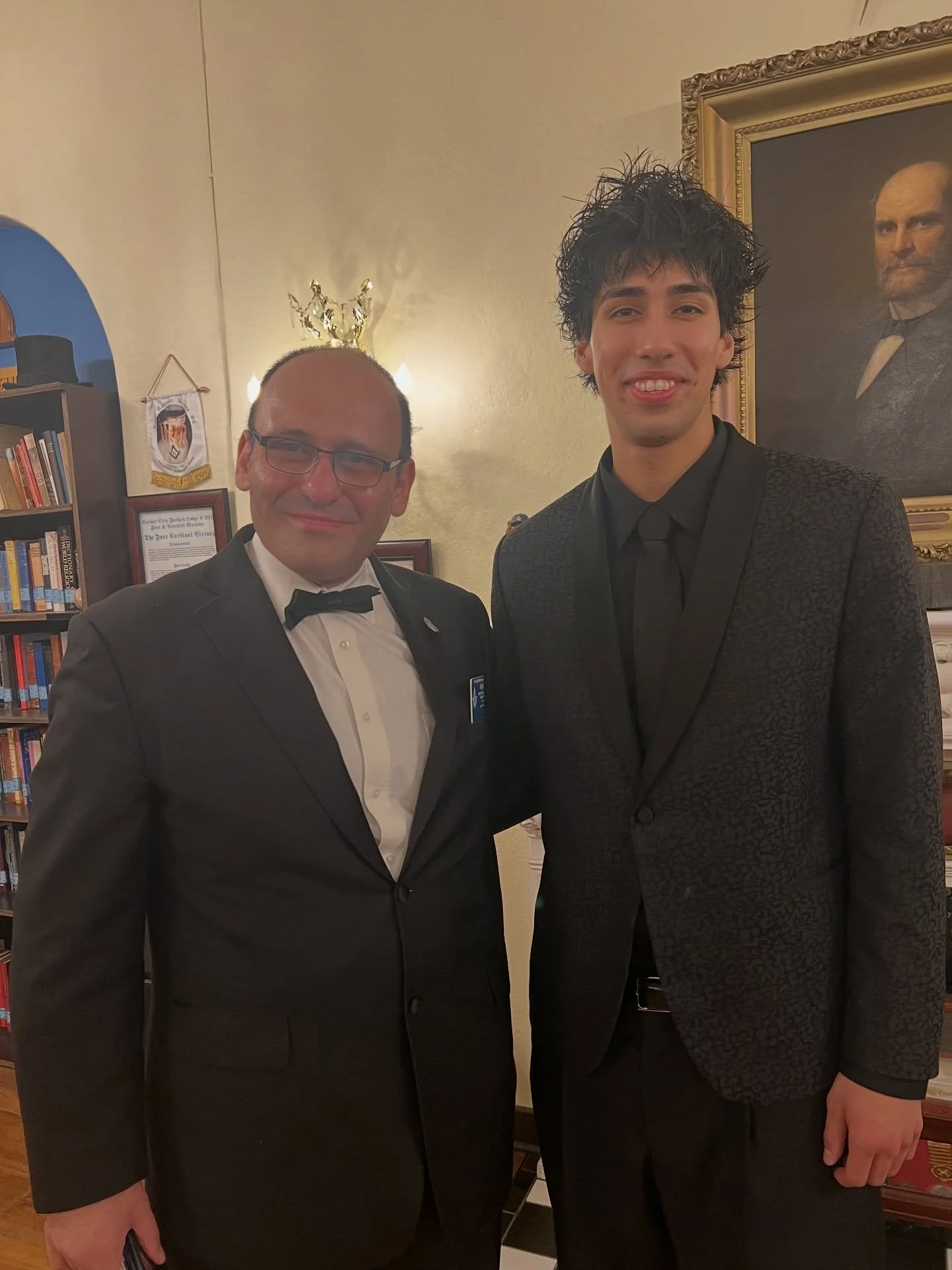  Cesar smiles beside a man in glasses and a bow tie, in front of a bookshelf and a framed portrait. 