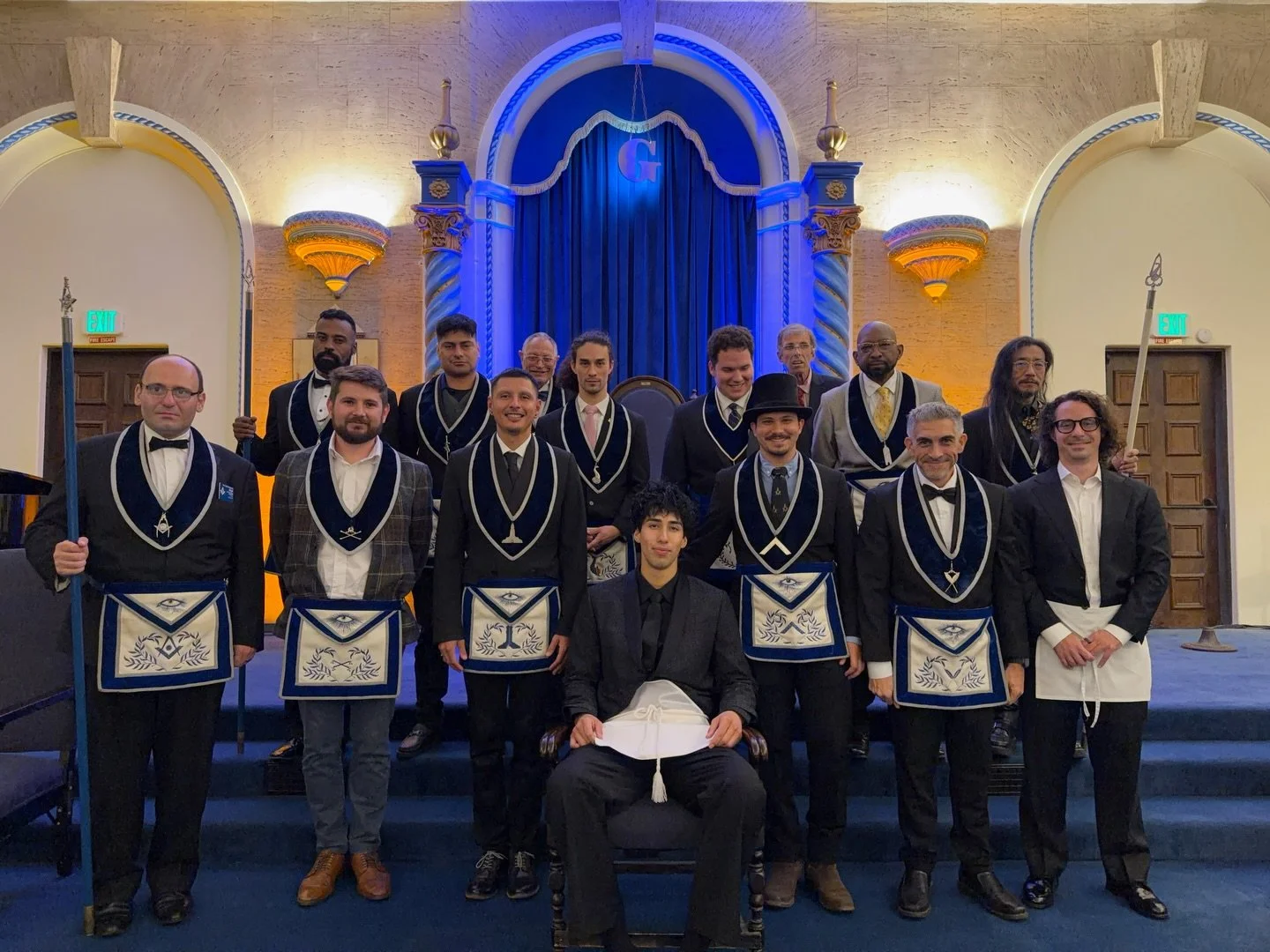  Cesar sits in front, holding a white apron, with about fifteen lodge brothers in blue regalia standing in the lodge room. 