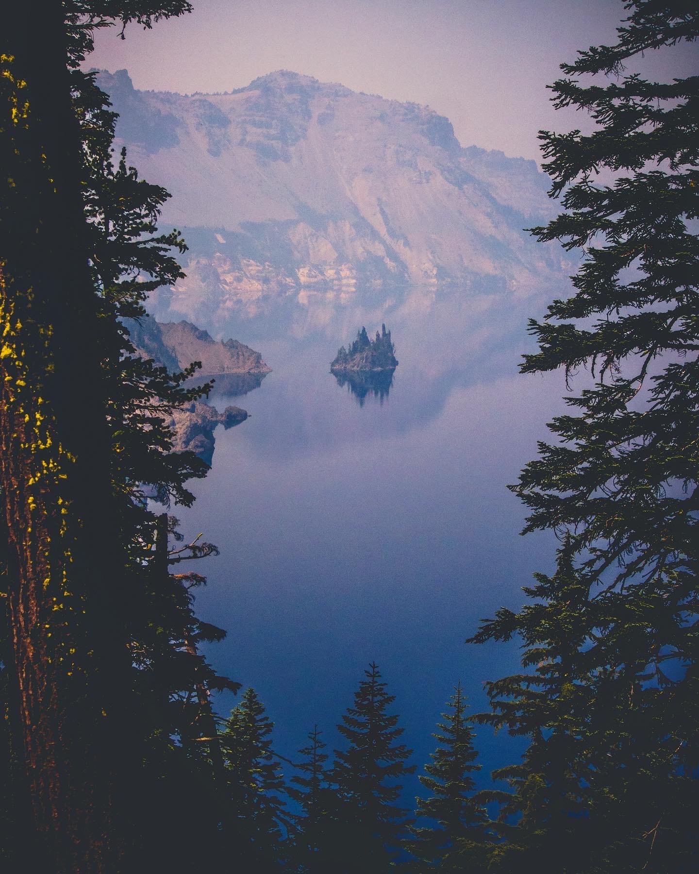 Misty mountain lake with rocky islands, framed by towering evergreen trees in Crater Lake National Park.