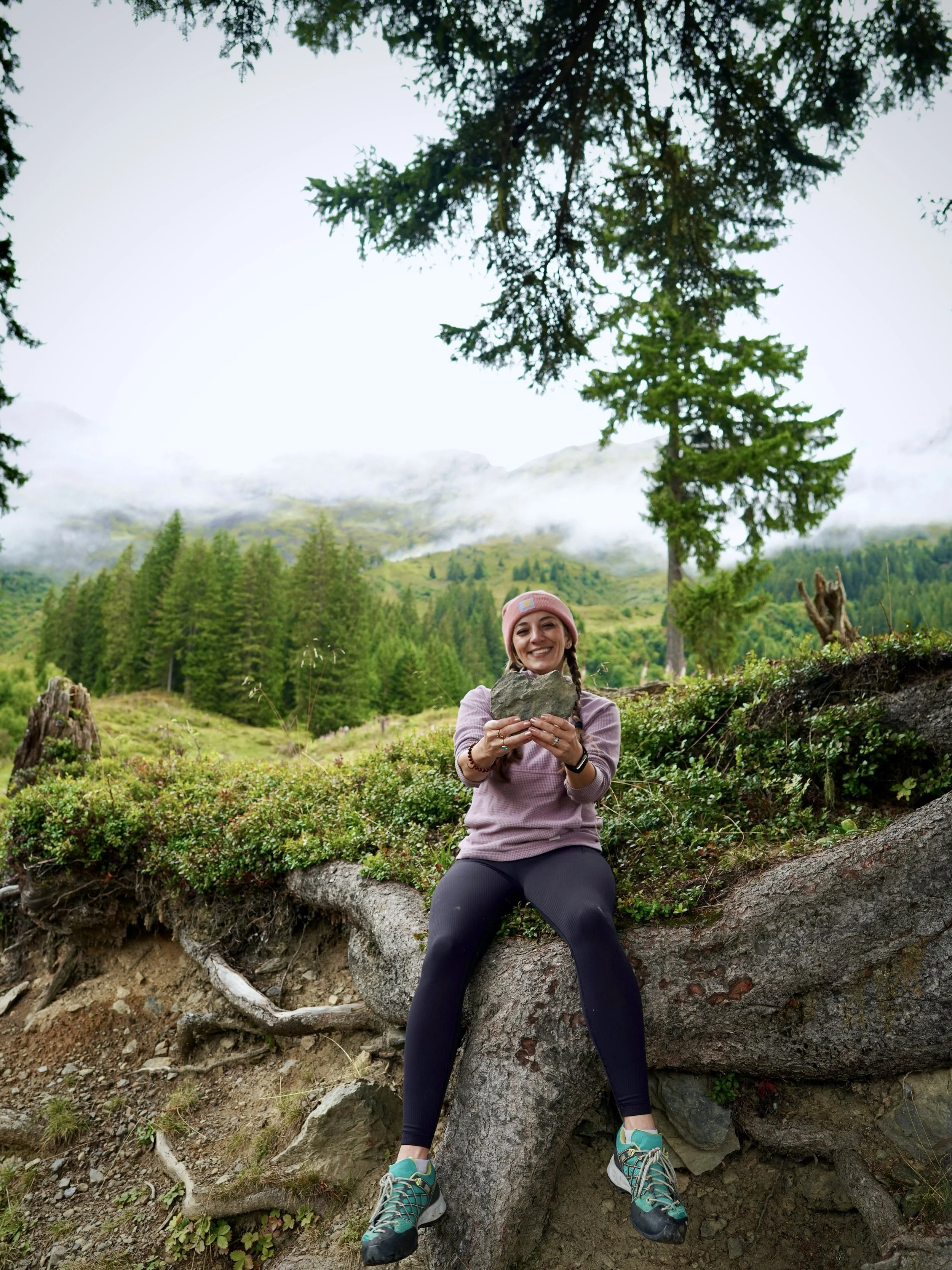 Melanie Boling in a pink beanie holding a rock while sitting on a tree trunk in a misty mountain forest in Murren, Switzerland.