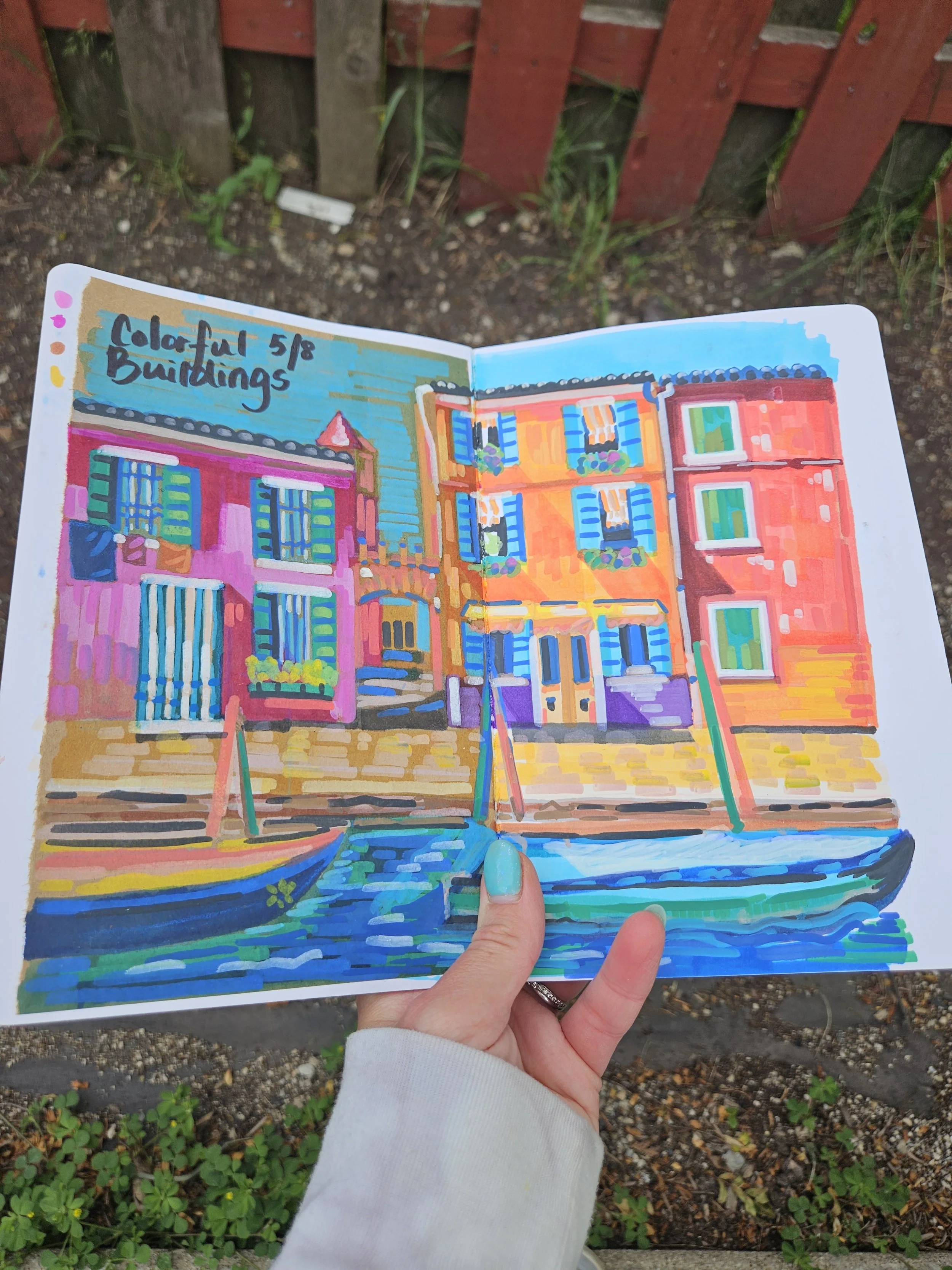 Colorful hand-painted drawing of row houses in a sketchbook held outdoors in front of a red fence.