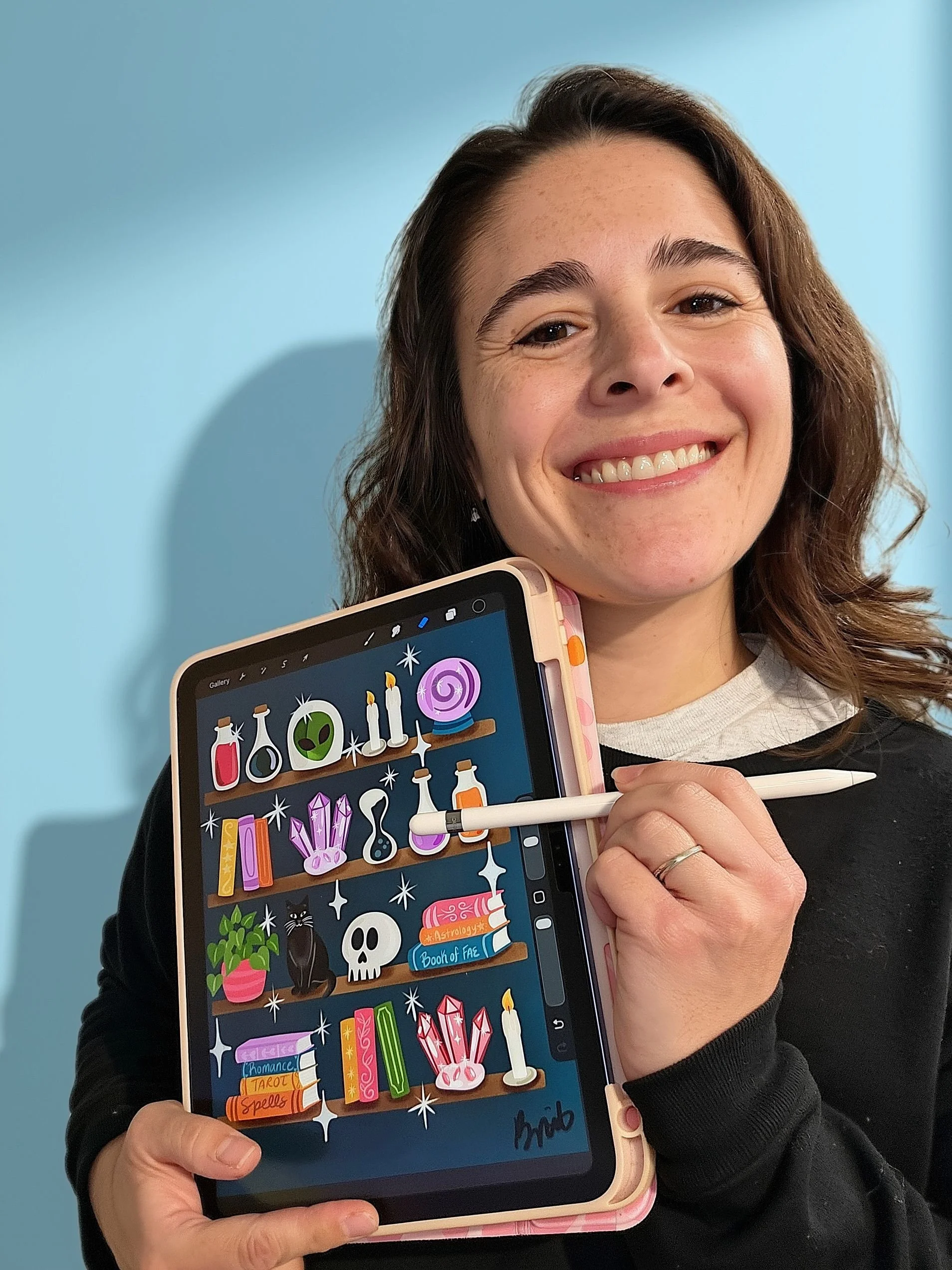 Smiling woman holding a tablet displaying colorful fantasy artwork of potion bottles, crystals, candles, spell books, and a black cat.