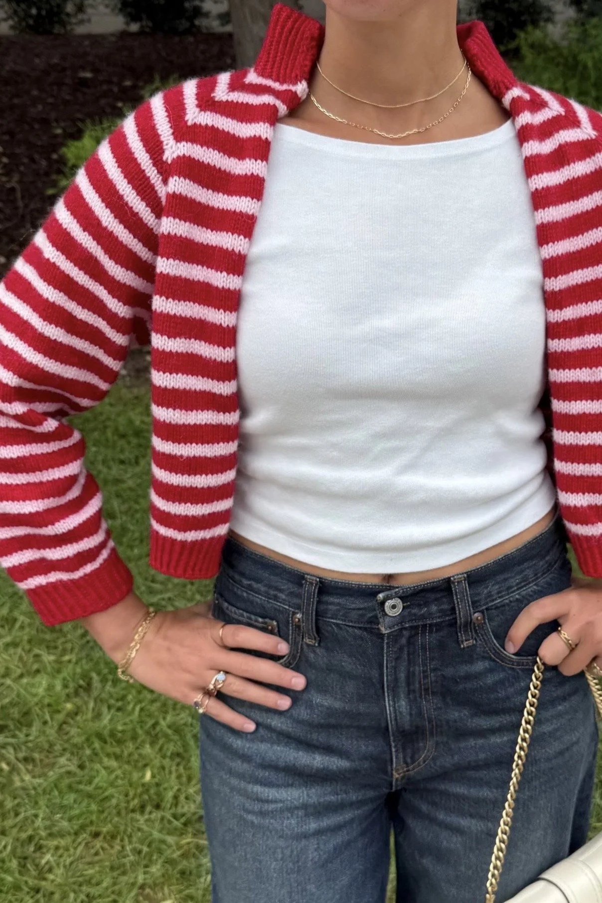 Person wearing a red-and-white striped knit cardigan, white tank top, dark jeans, layered gold necklaces, and jewelry outdoors.
