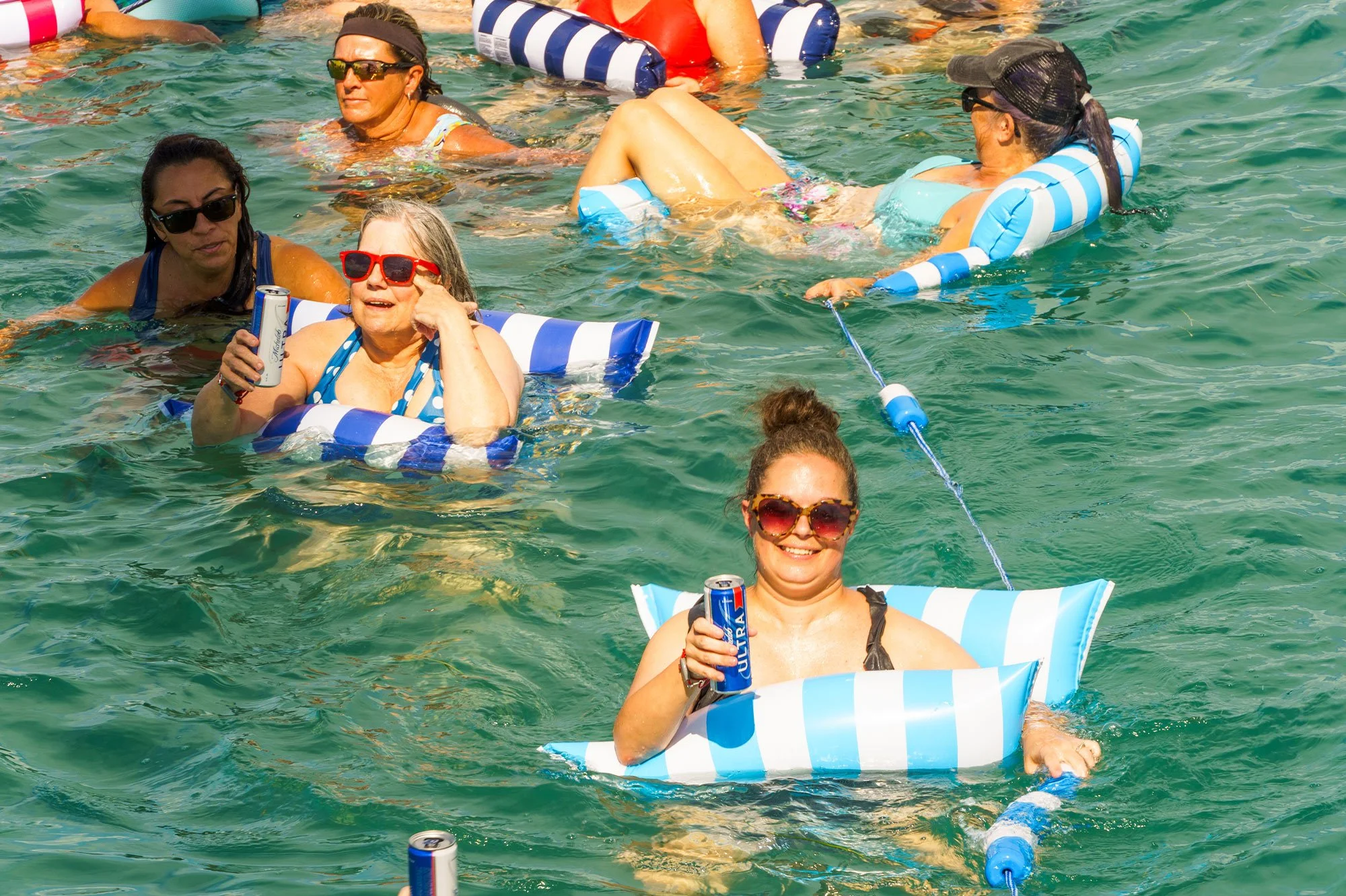 Girls Gone Cruisin' group laughing on striped pool floats in turquoise water