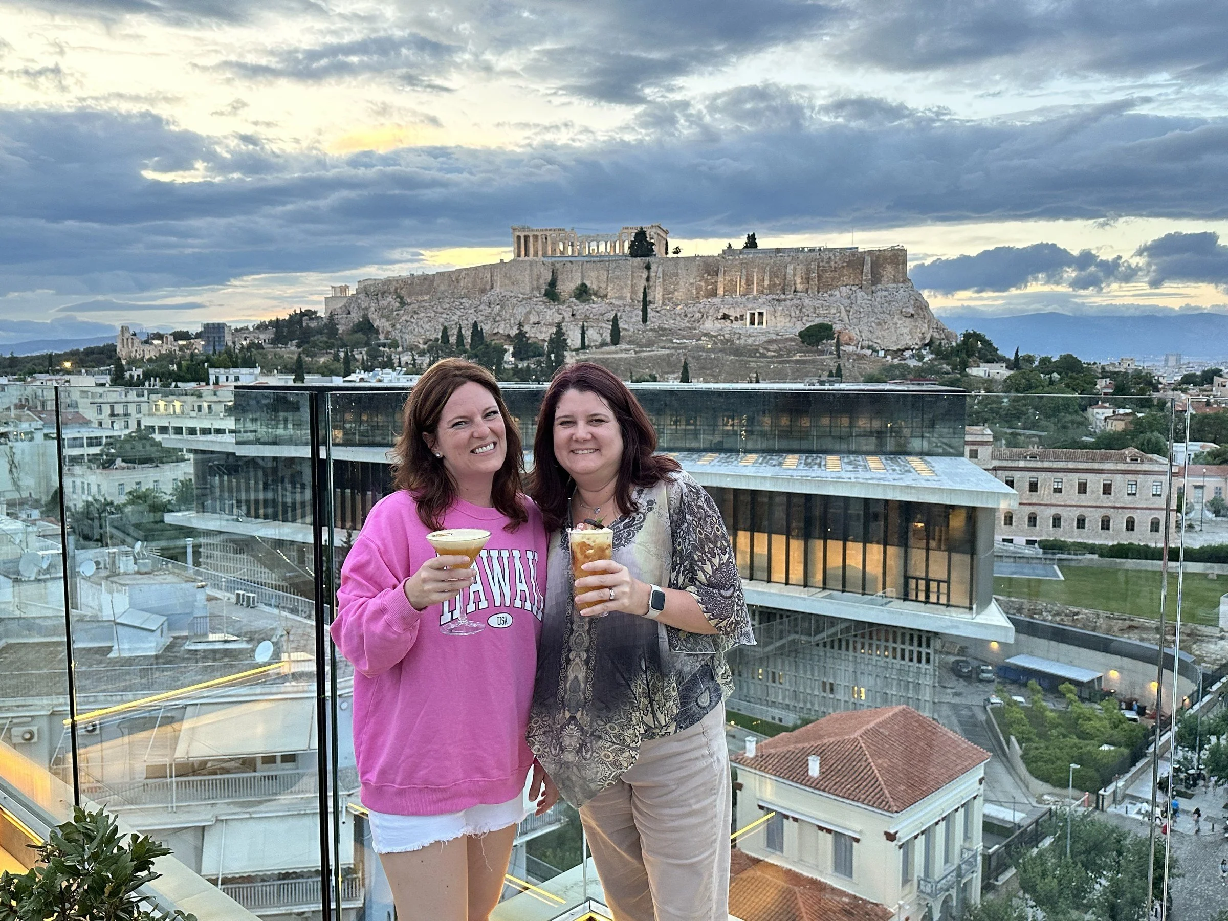 Samantha and a friend toasting with drinks on a rooftop terrace in Athens, with the Acropolis on the hill behind them