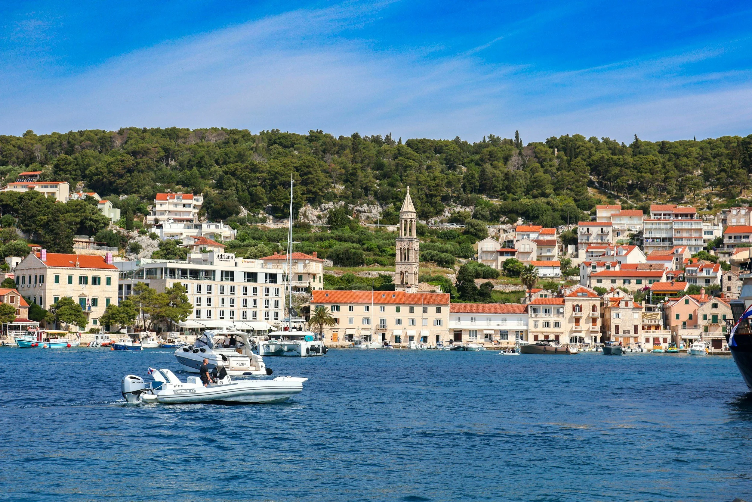 Hvar town's waterfront, with a bell tower and stone buildings rising up a green hillside and a motorboat in the harbor