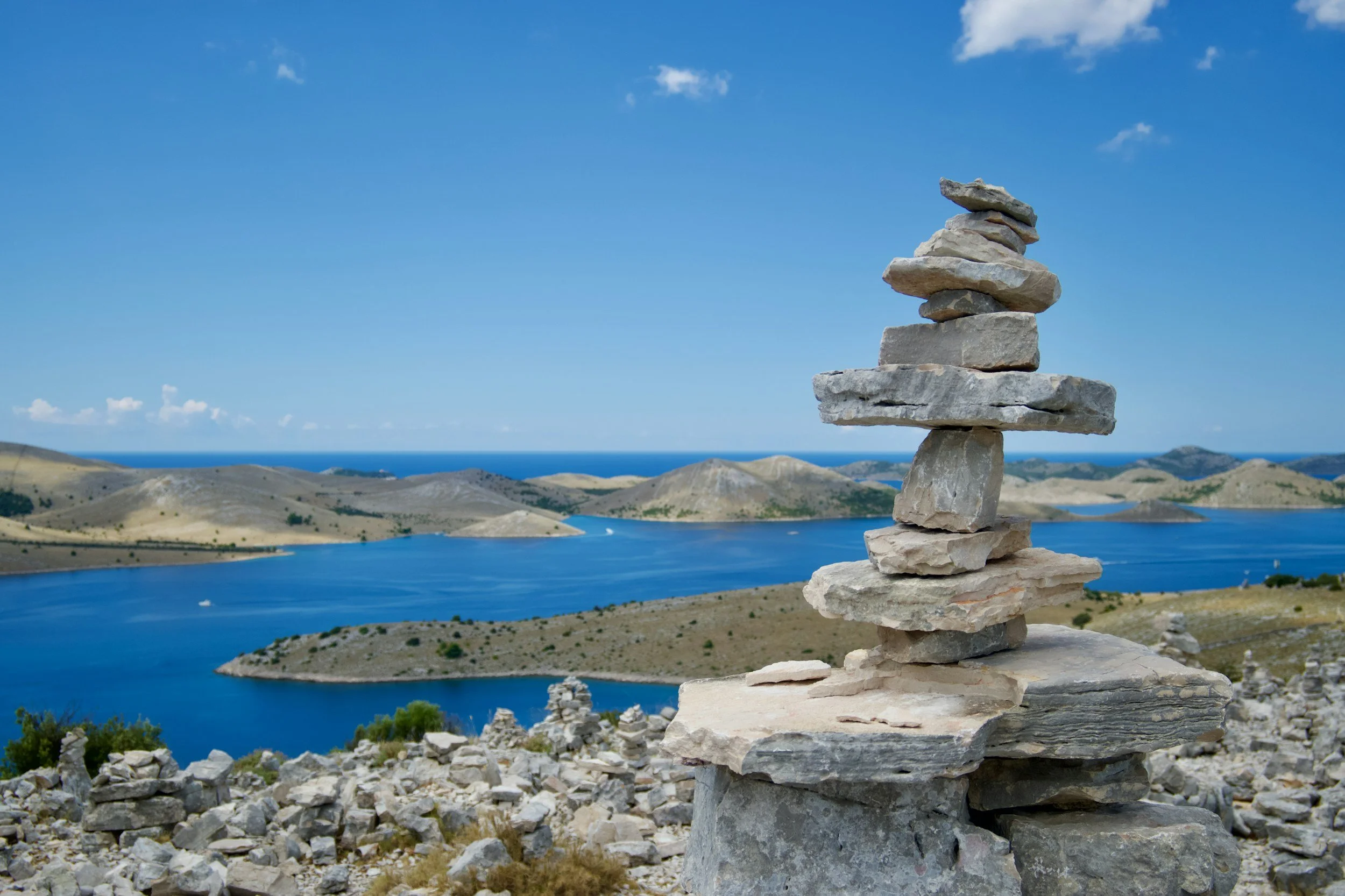 A stacked-stone cairn on a rocky hilltop overlooking low, bare islands scattered across a deep blue sea