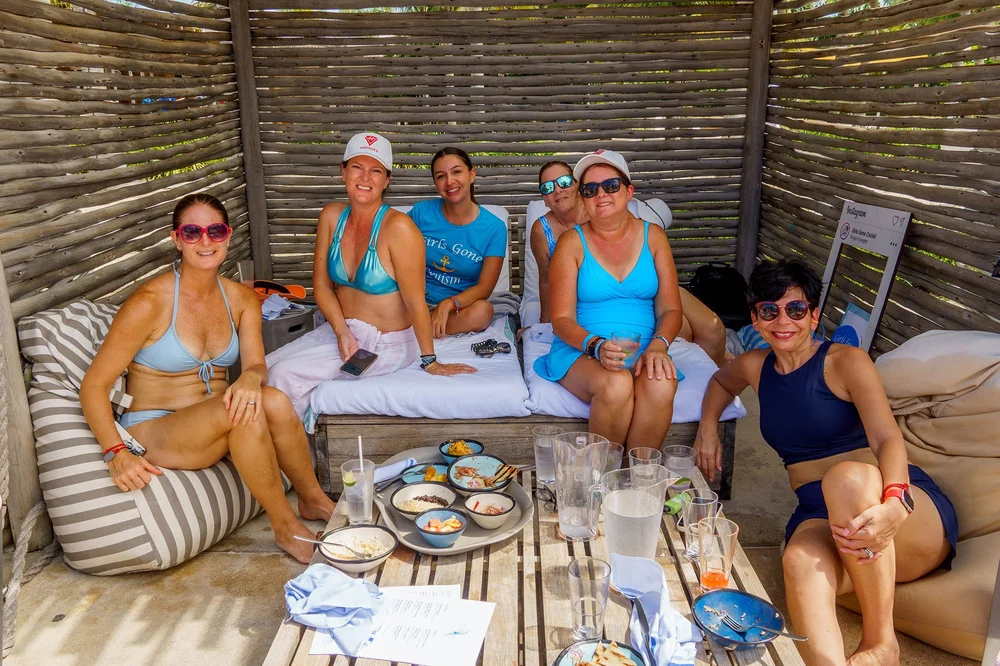 Women in swimsuits sharing food in a shaded beach cabana