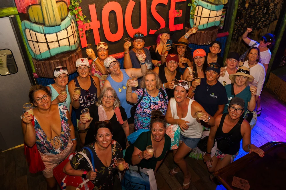 A crowd of women raising drinks under a neon House sign at a bar